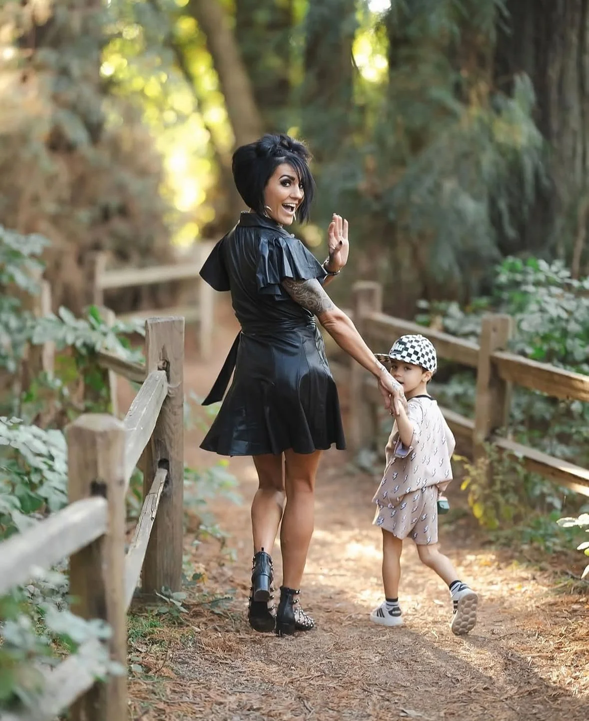 A woman and a young boy walking along a wooded path, holding hands, the woman smiling and waving, with sunlight filtering through trees in the background.