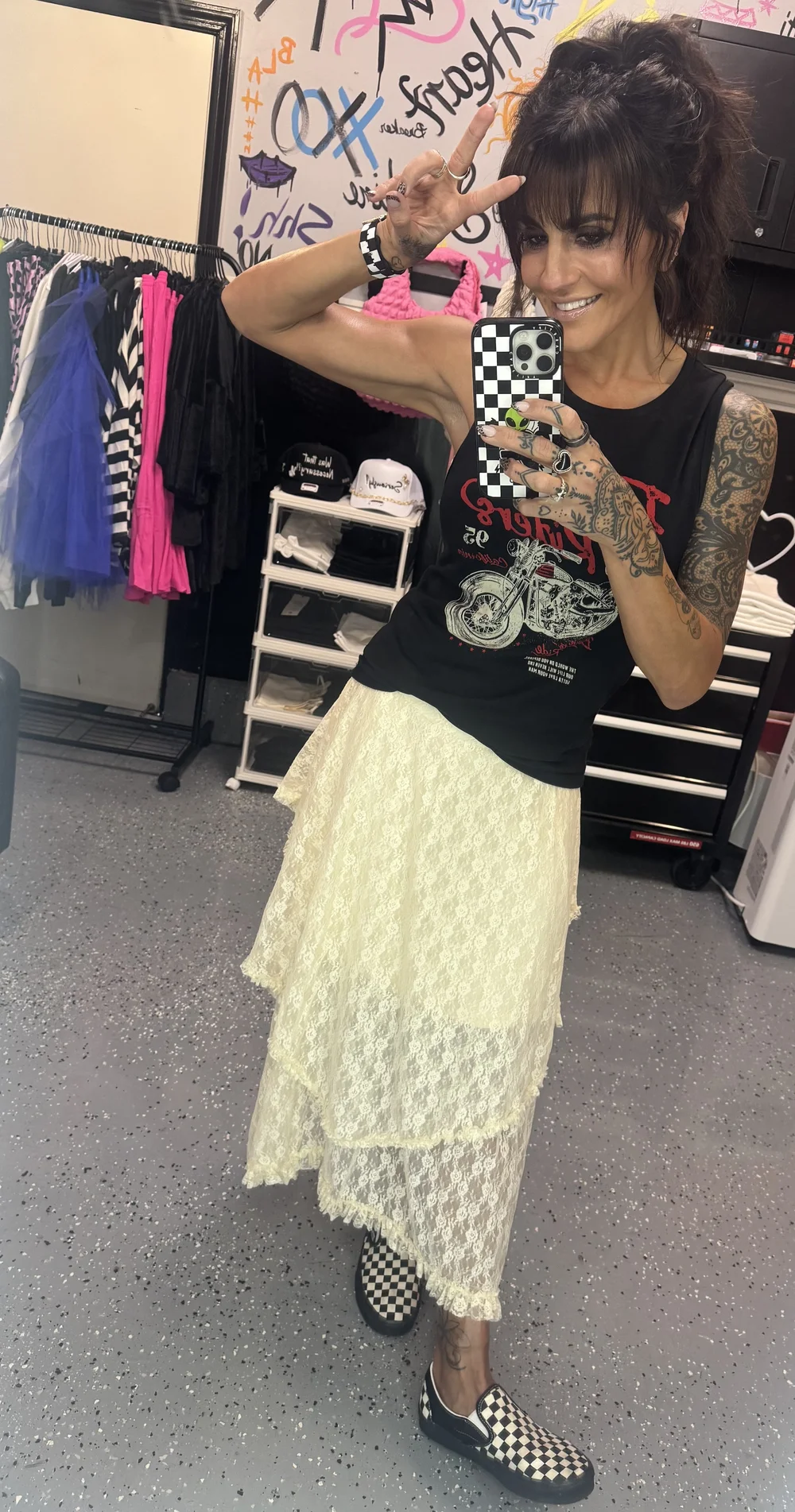Cari in the shop wearing a band tank, yellow lace skirt, and checkerboard slip-ons