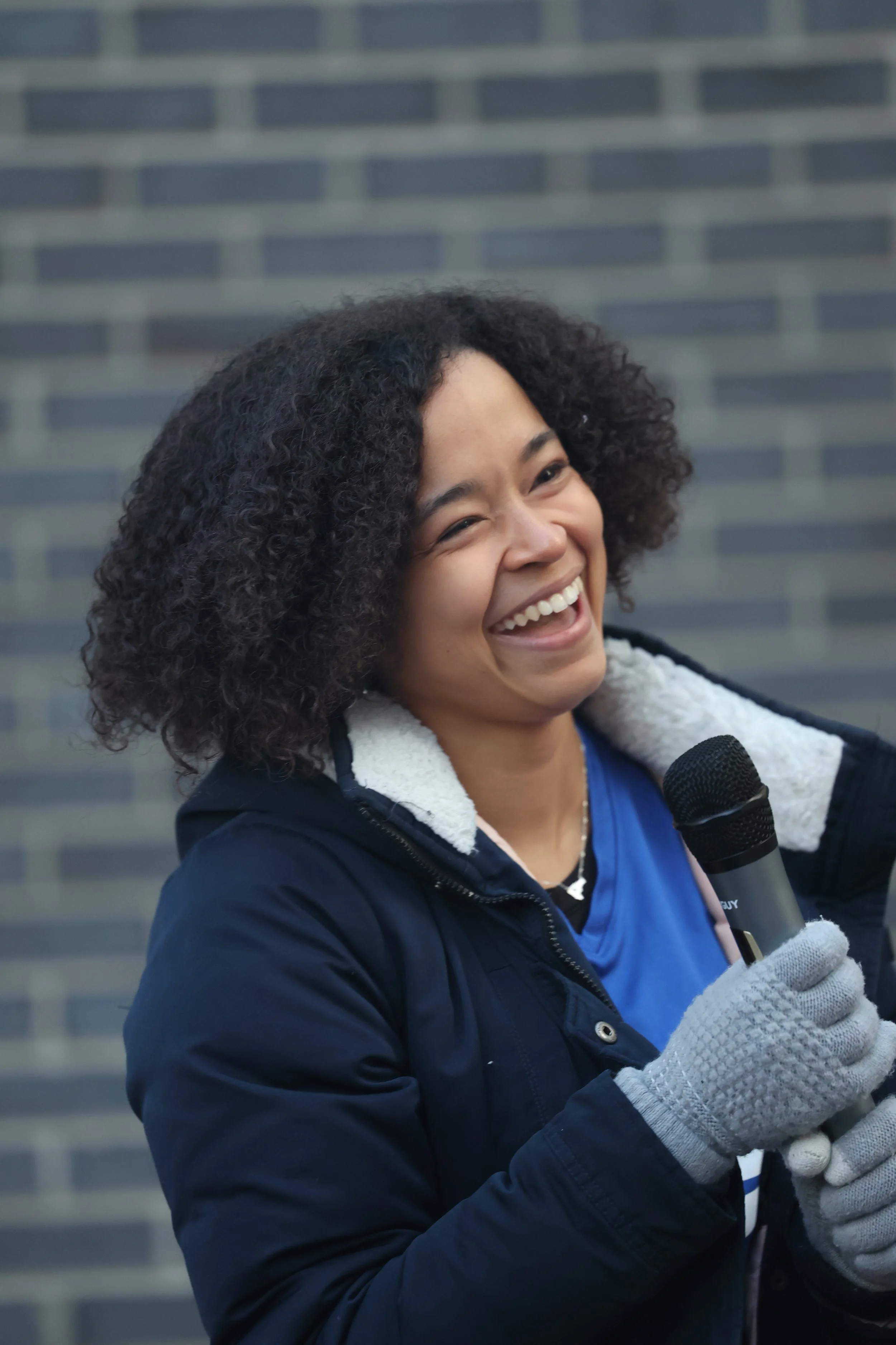 A woman with curly hair smiling and laughing while holding a microphone, wearing a dark jacket and gloves, standing in front of a brick wall.