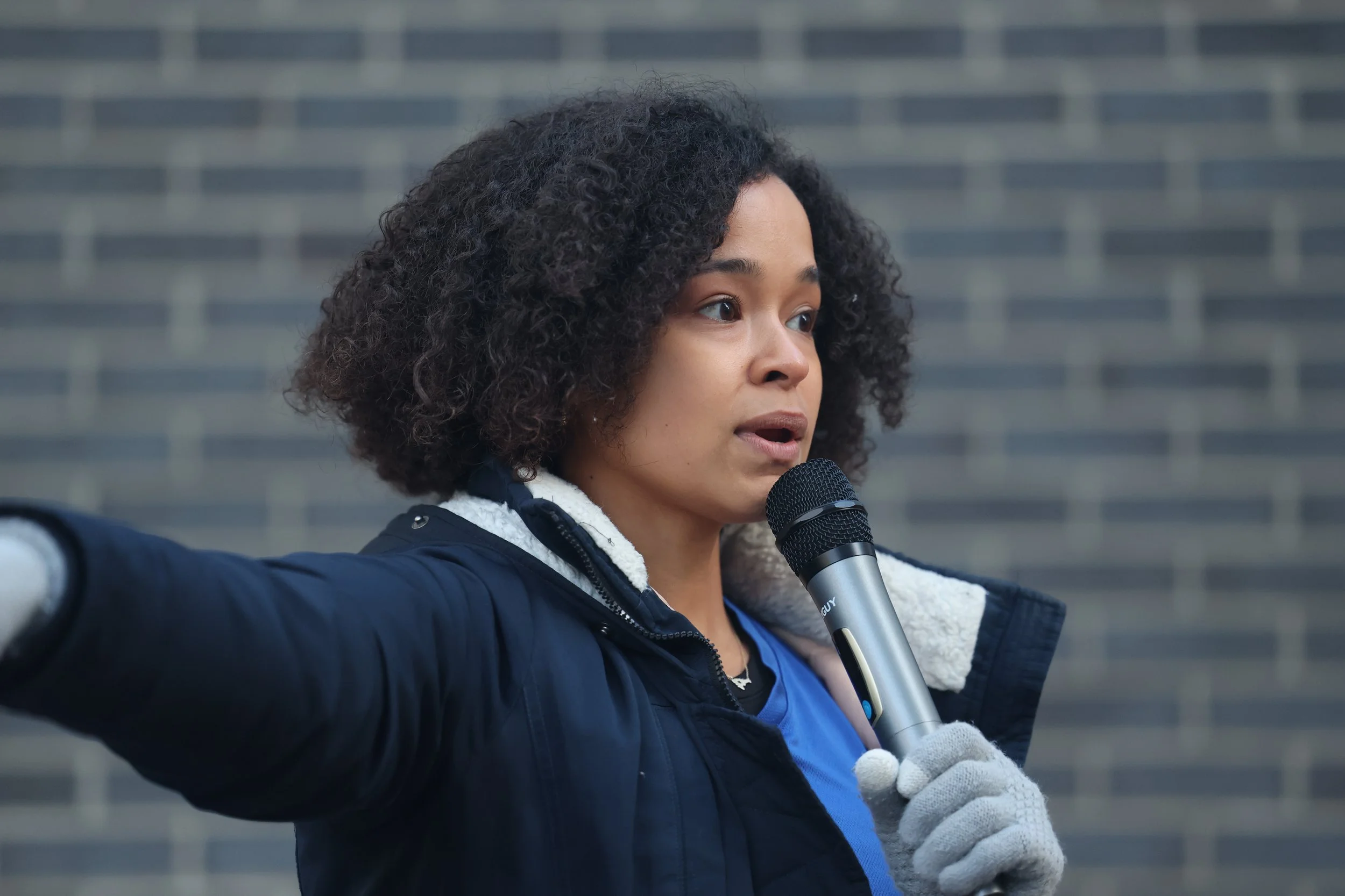 A woman with curly hair speaking into a microphone outdoors, wearing a navy jacket with a fleece-lined hood and white gloves, with a blurred brick wall in the background.