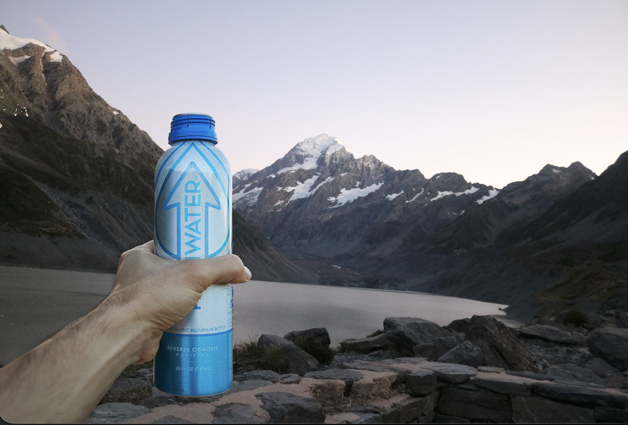 A person holding a blue aluminum water bottle with mountains and a lake in the background during sunset or sunrise.