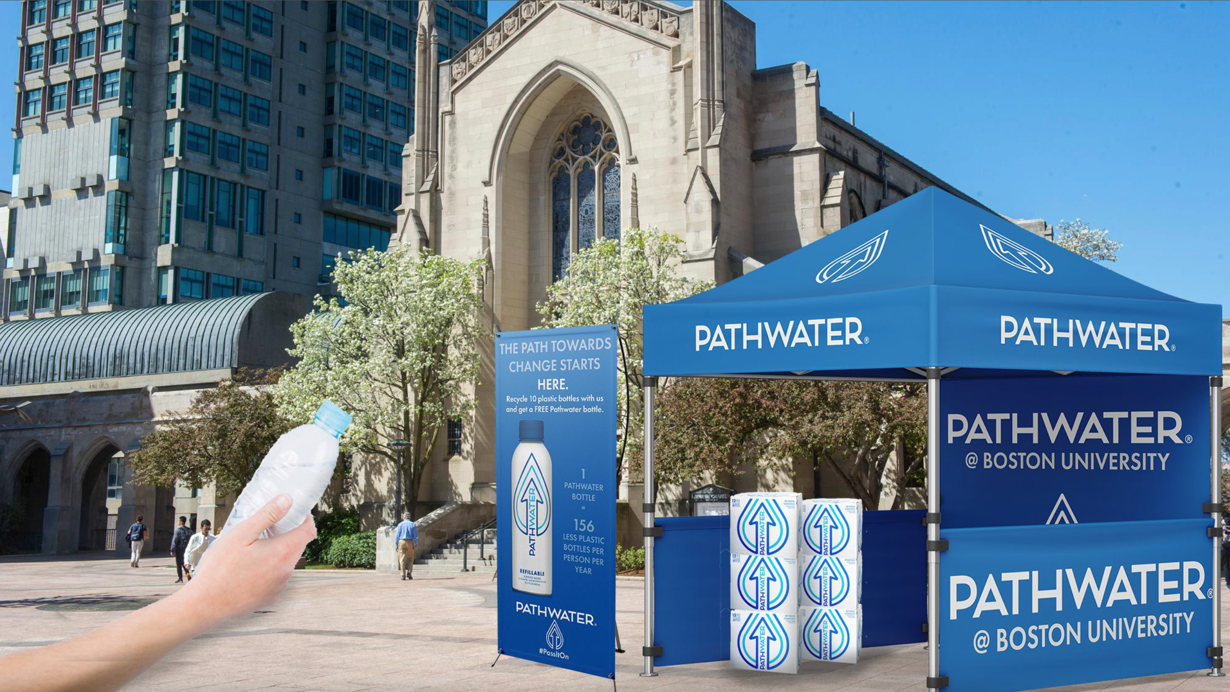 A Pathwater stand at Boston University with bottled water, banners, and a tent, in an outdoor campus area with trees and a church building with stained glass windows in the background. A hand holding a plastic water bottle is visible in the foreground.