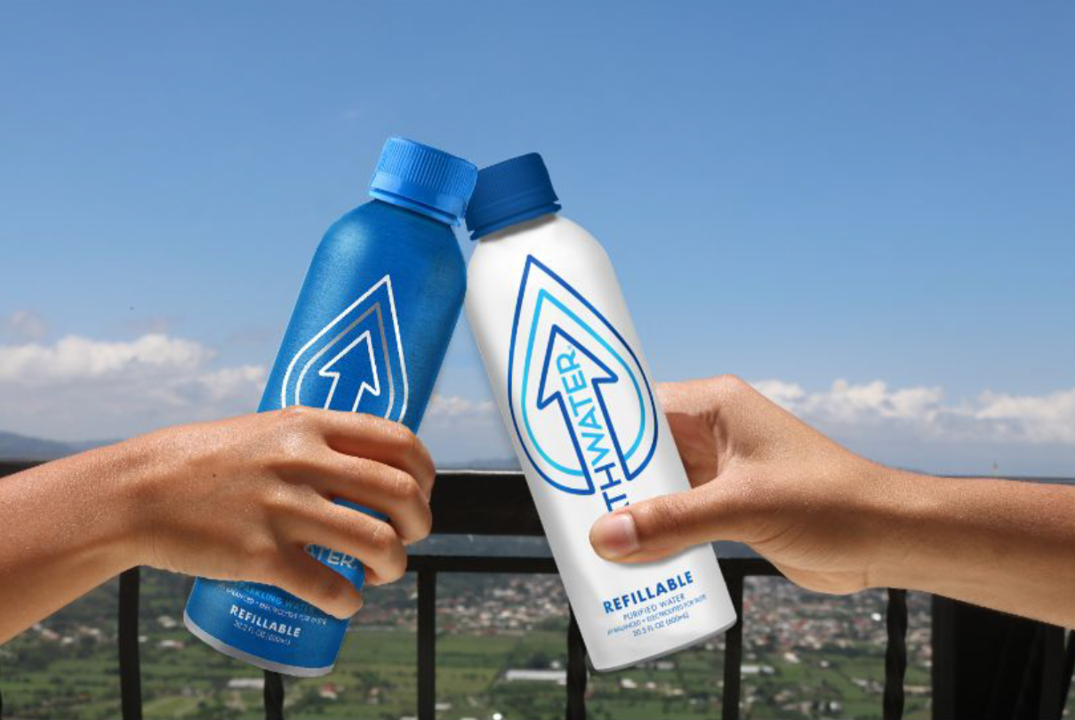 Two hands holding reusable water bottles in a toast with a mountain landscape and blue sky in the background.