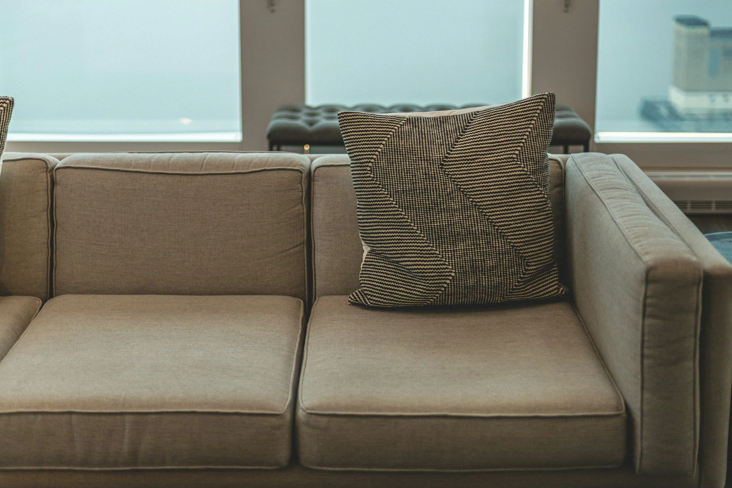 A beige sectional sofa with a chevron-patterned throw pillow in a room with large windows and a bench in the background.