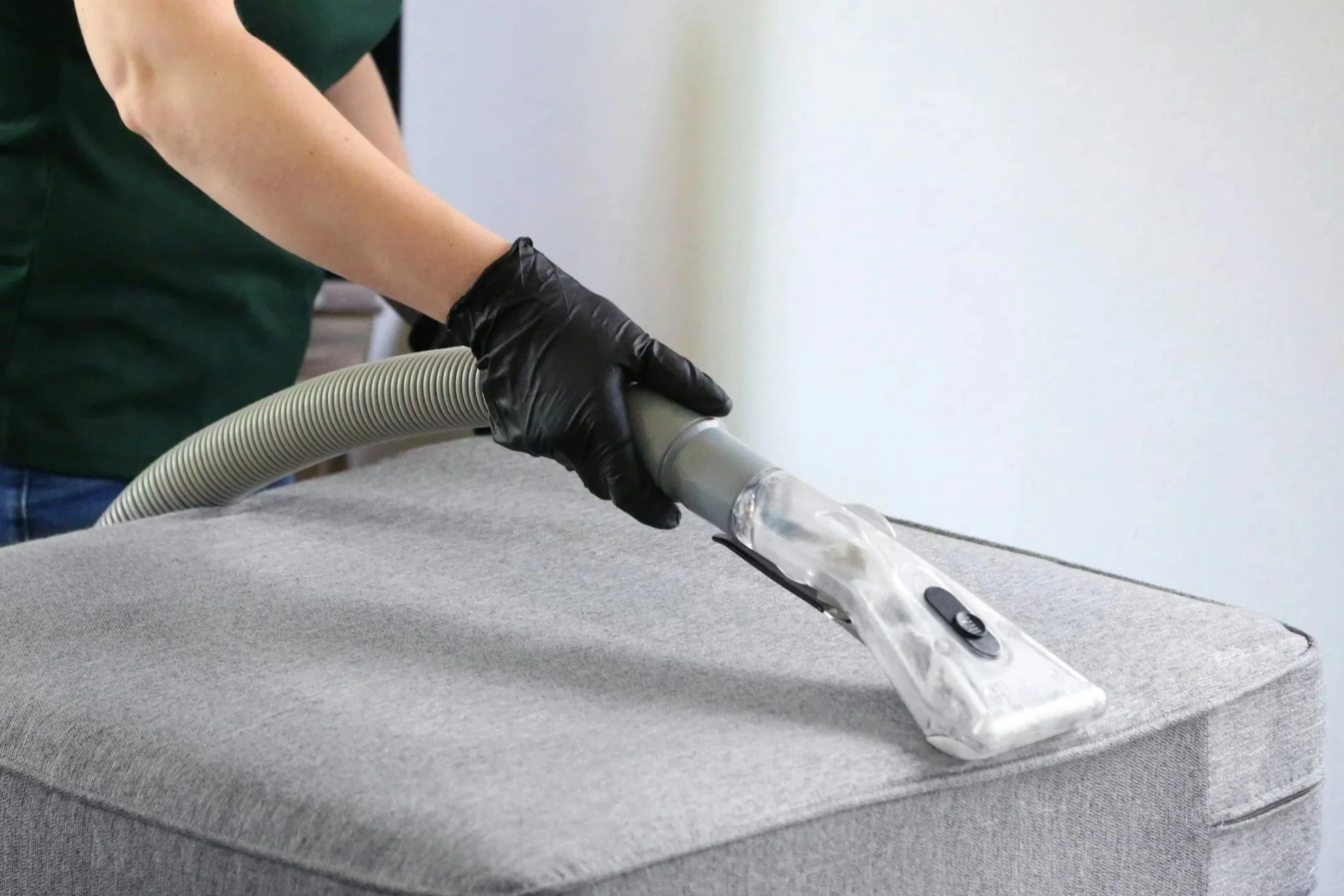 Person wearing black gloves vacuuming a gray upholstered furniture with a clear vacuum tube.