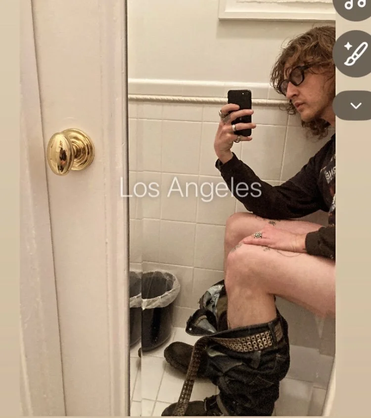 A person sitting on a toilet in a bathroom, taking a mirror selfie, with the text 'Los Angeles' overlayed on the image.