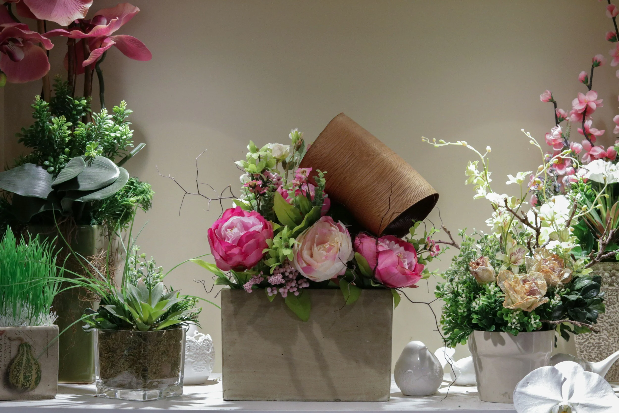A display of various potted plants and flowers, including pink peonies, succulents, and greenery, arranged on a table with a plain background.