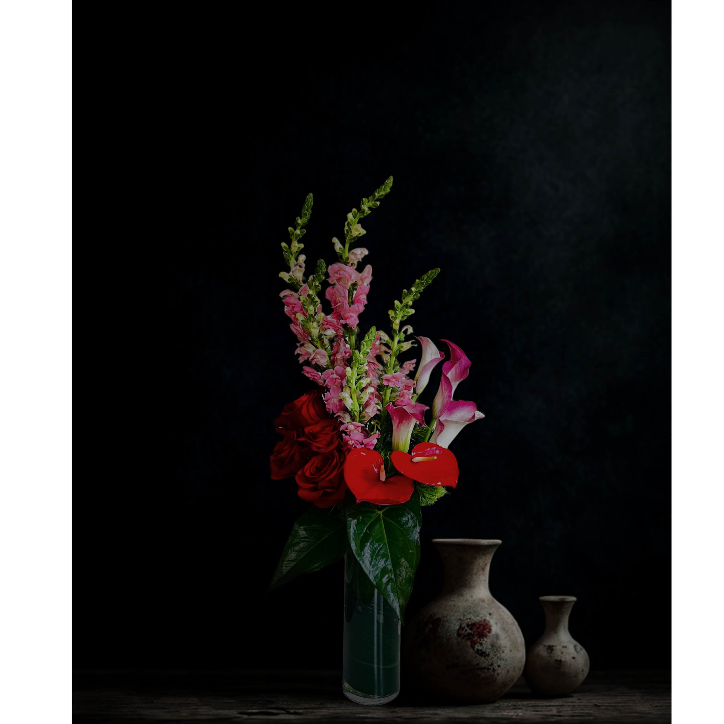 A floral arrangement in a green glass vase on a dark wooden surface, with a textured black background and decorative stone objects in the background.