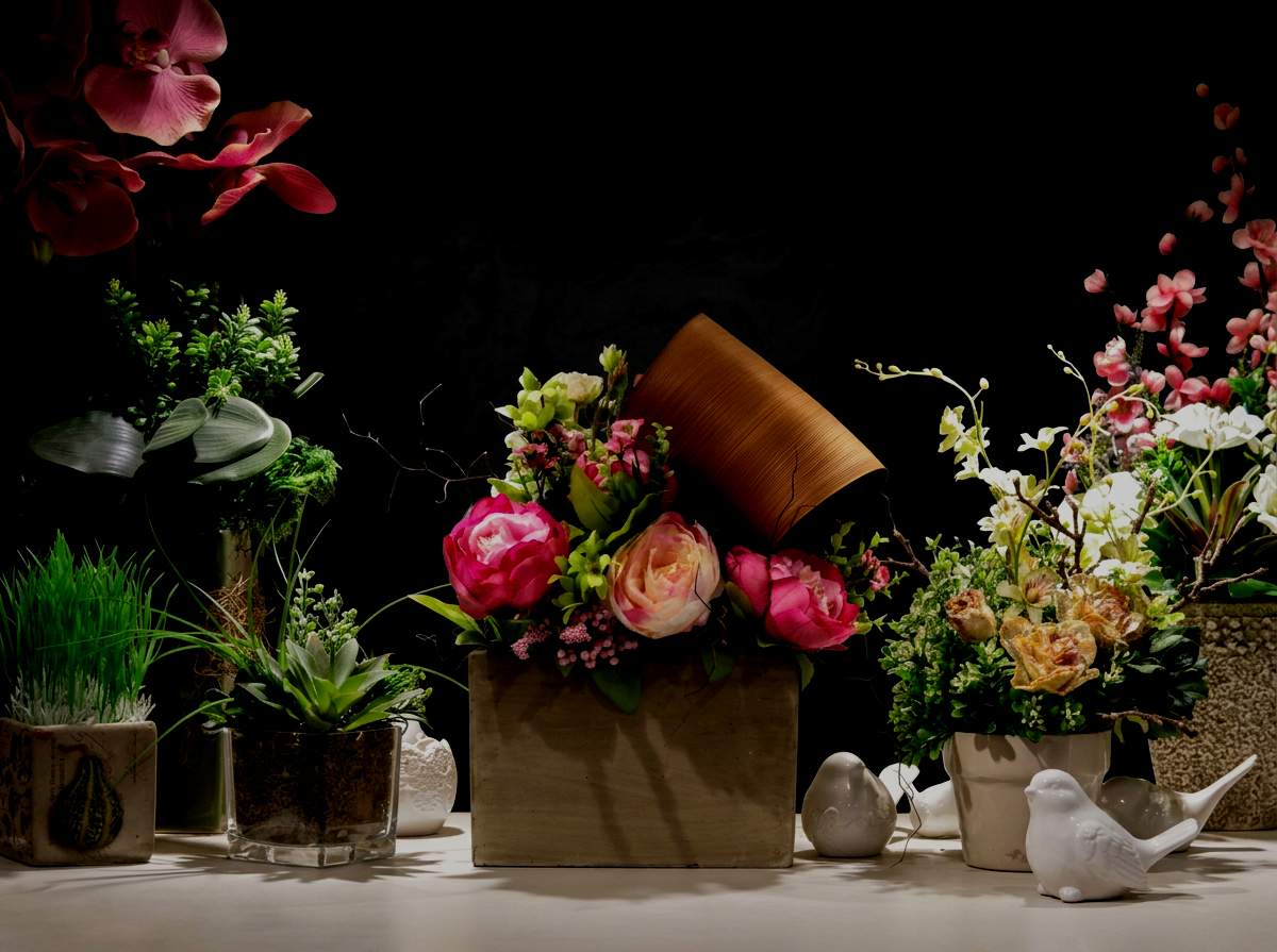 Various potted plants and flowers arranged on a table, including pink, white, and peach roses, orchids, and green foliage, with decorative bird figurines and a dark background.