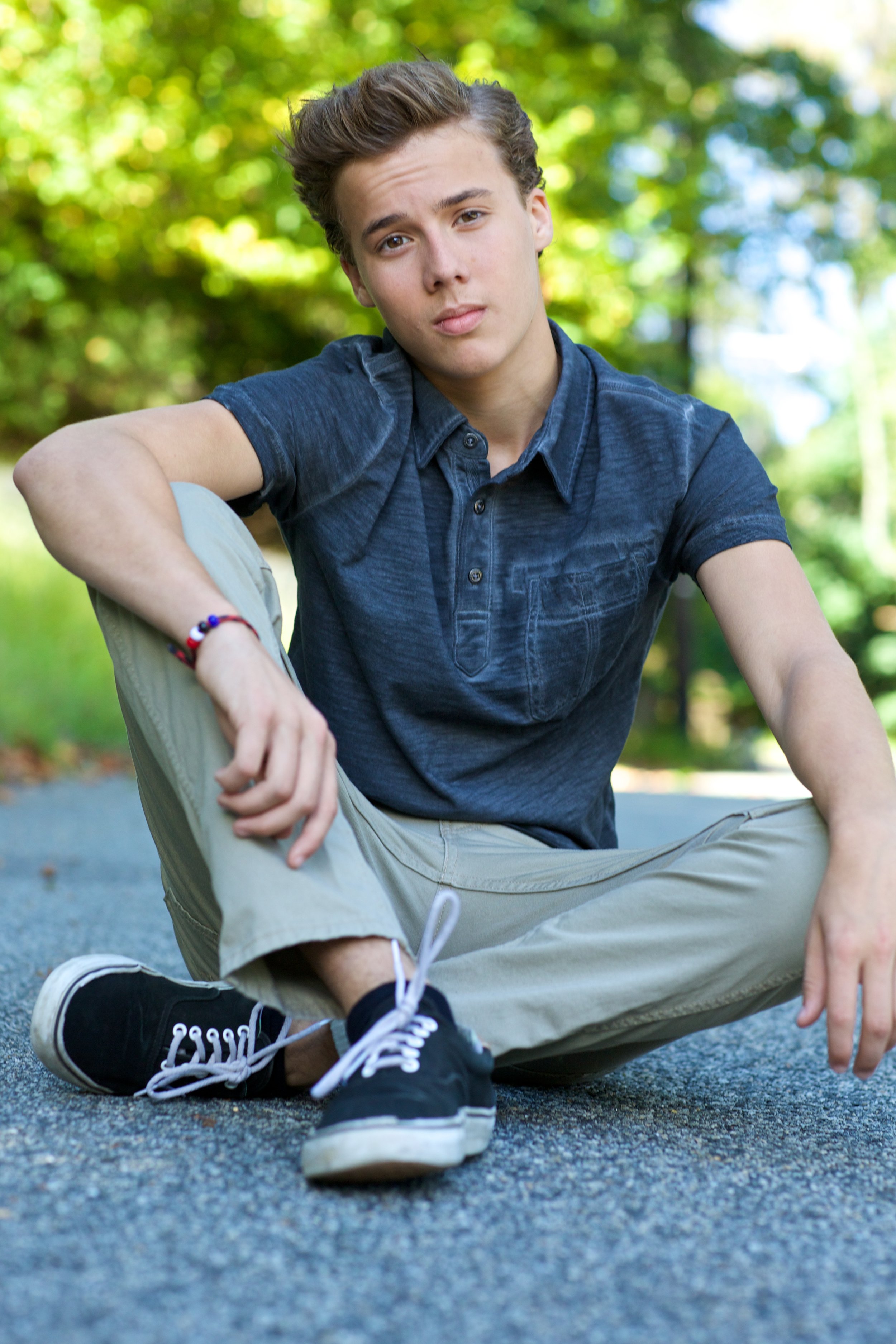 A young man with short brown hair sitting on a paved surface outdoors, wearing a navy blue button-up shirt, khaki pants, black sneakers, and a red and blue bracelet, with a background of green, blurred trees.