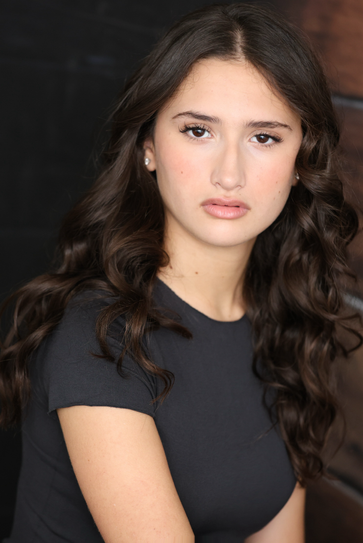 A young woman with long, dark wavy hair and light makeup, wearing a black shirt, looking directly at the camera with a neutral expression.