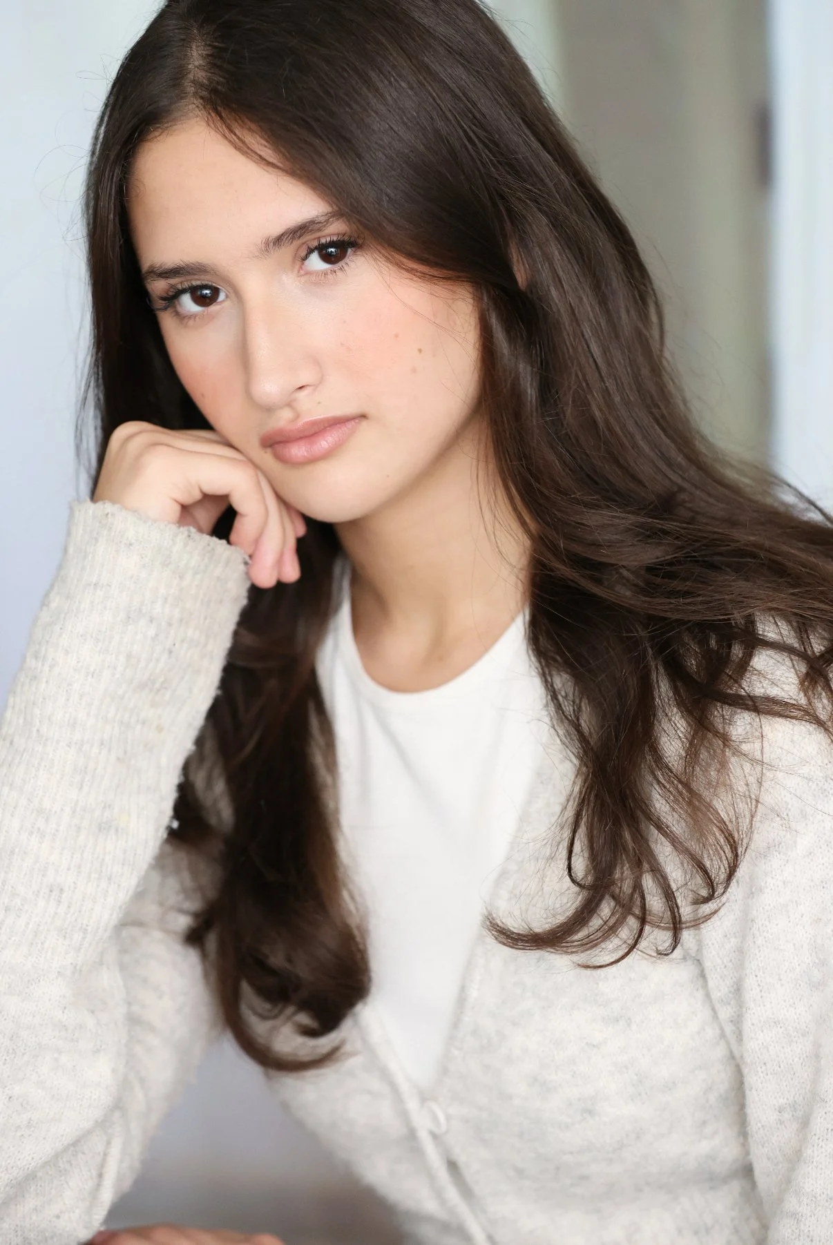 A young woman with long, wavy brown hair, wearing a white top and beige cardigan, resting her chin on her hand and looking at the camera.