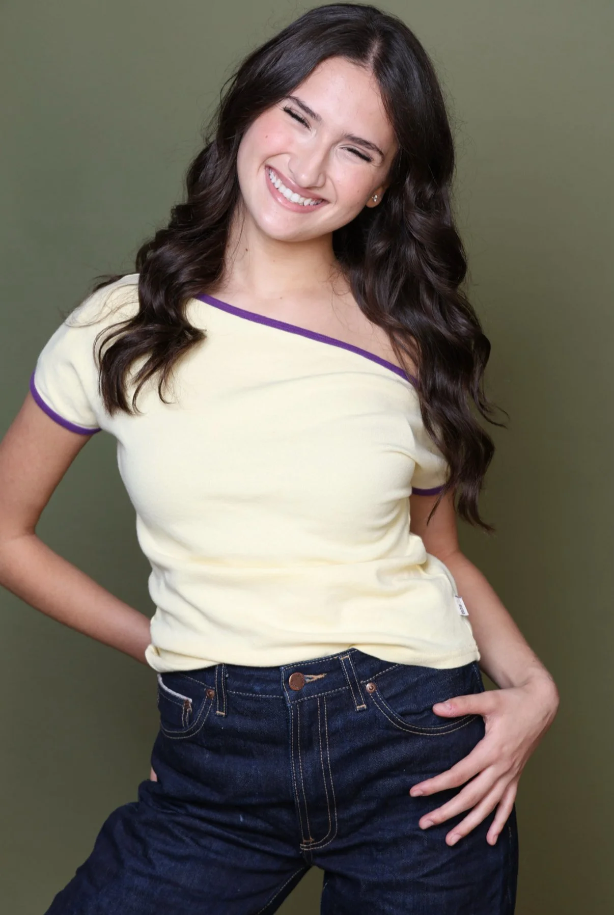 Young woman with long dark hair, smiling with eyes closed, wearing a yellow top with purple trim and dark jeans, standing against a green background.