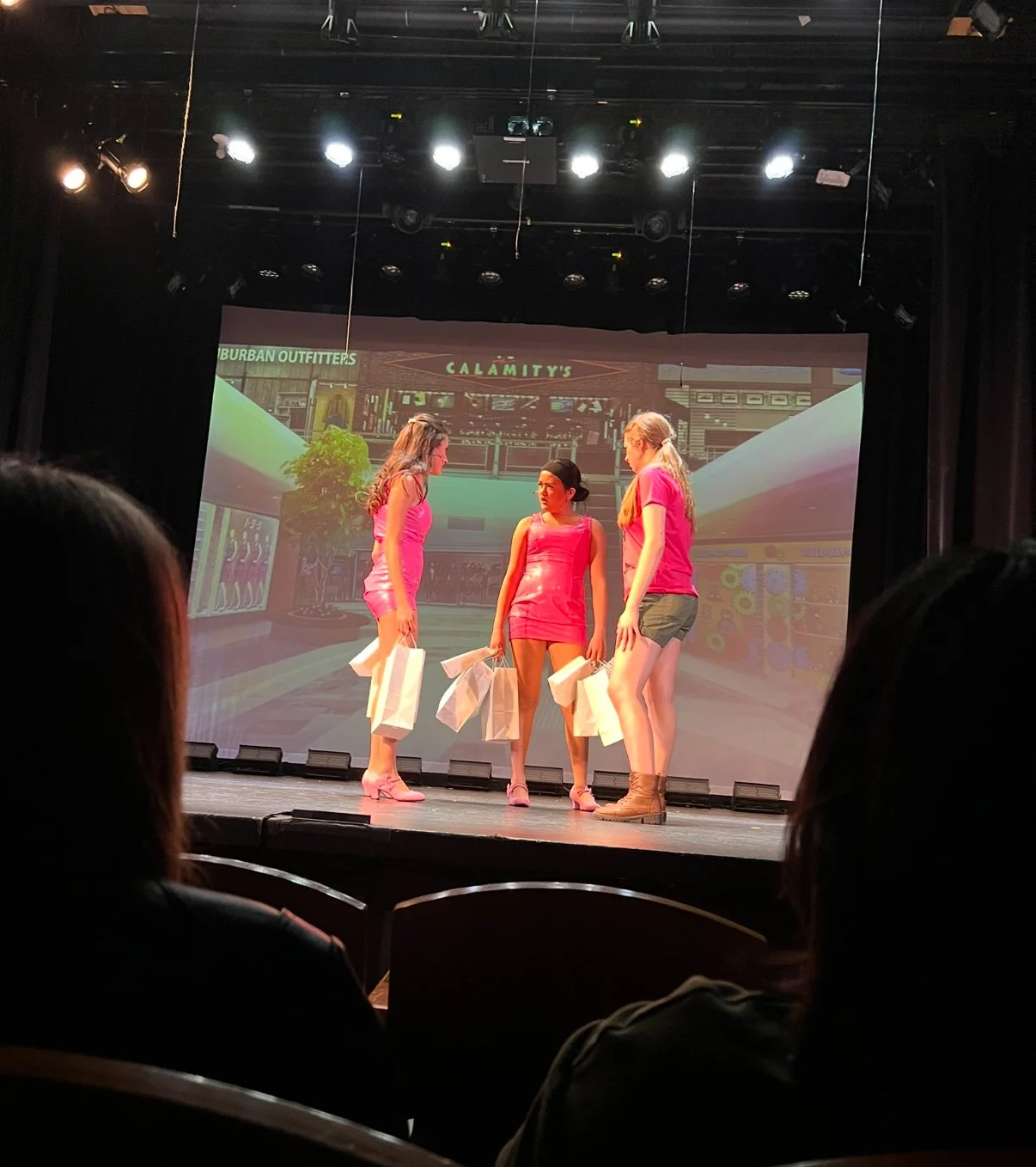 Four women on stage in pink outfits, holding shopping bags, with a cityscape background projected behind them during a performance or presentation.
