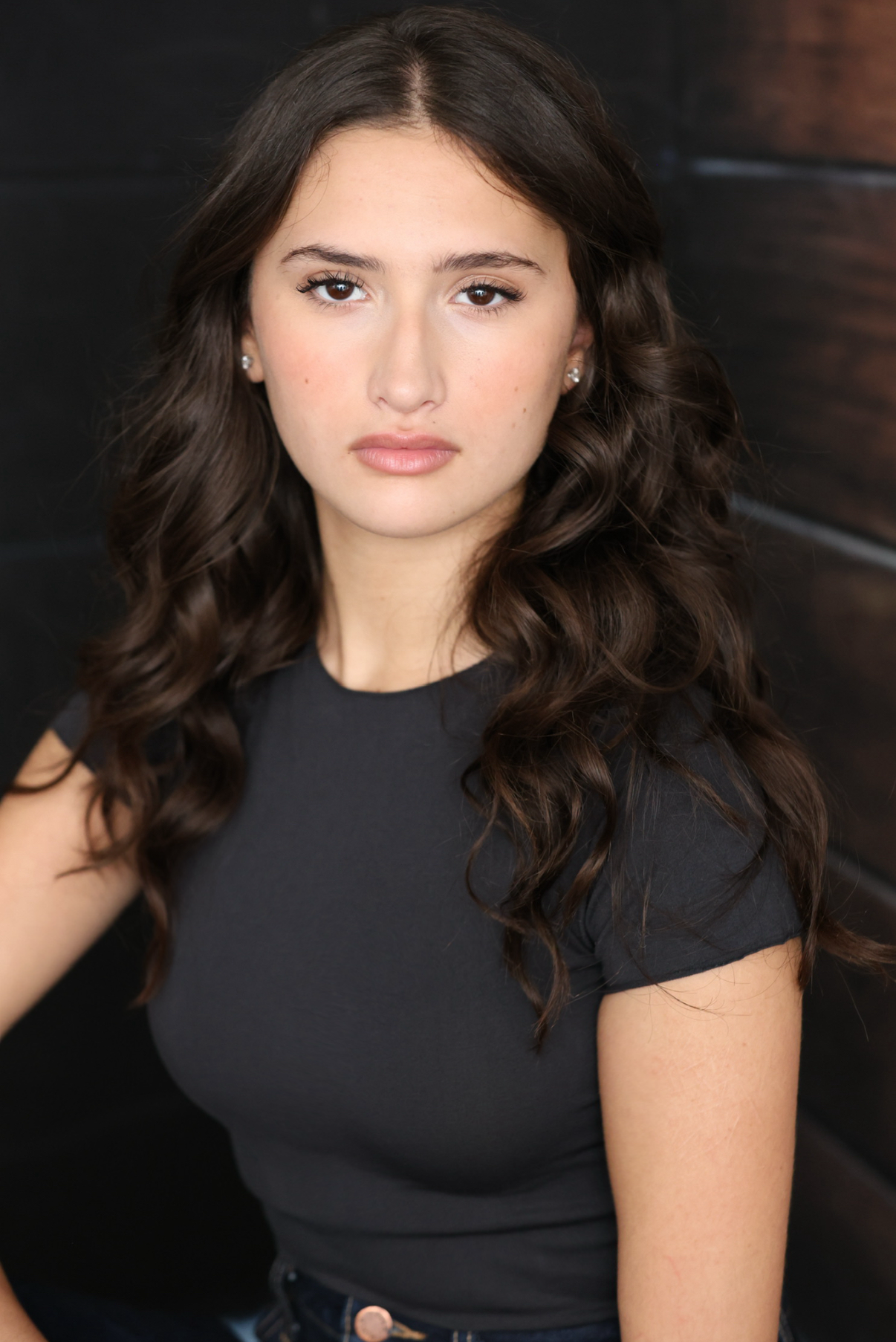 A young woman with long, wavy brown hair, wearing a black t-shirt, looking directly at the camera with a neutral expression, standing against a dark wooden background.