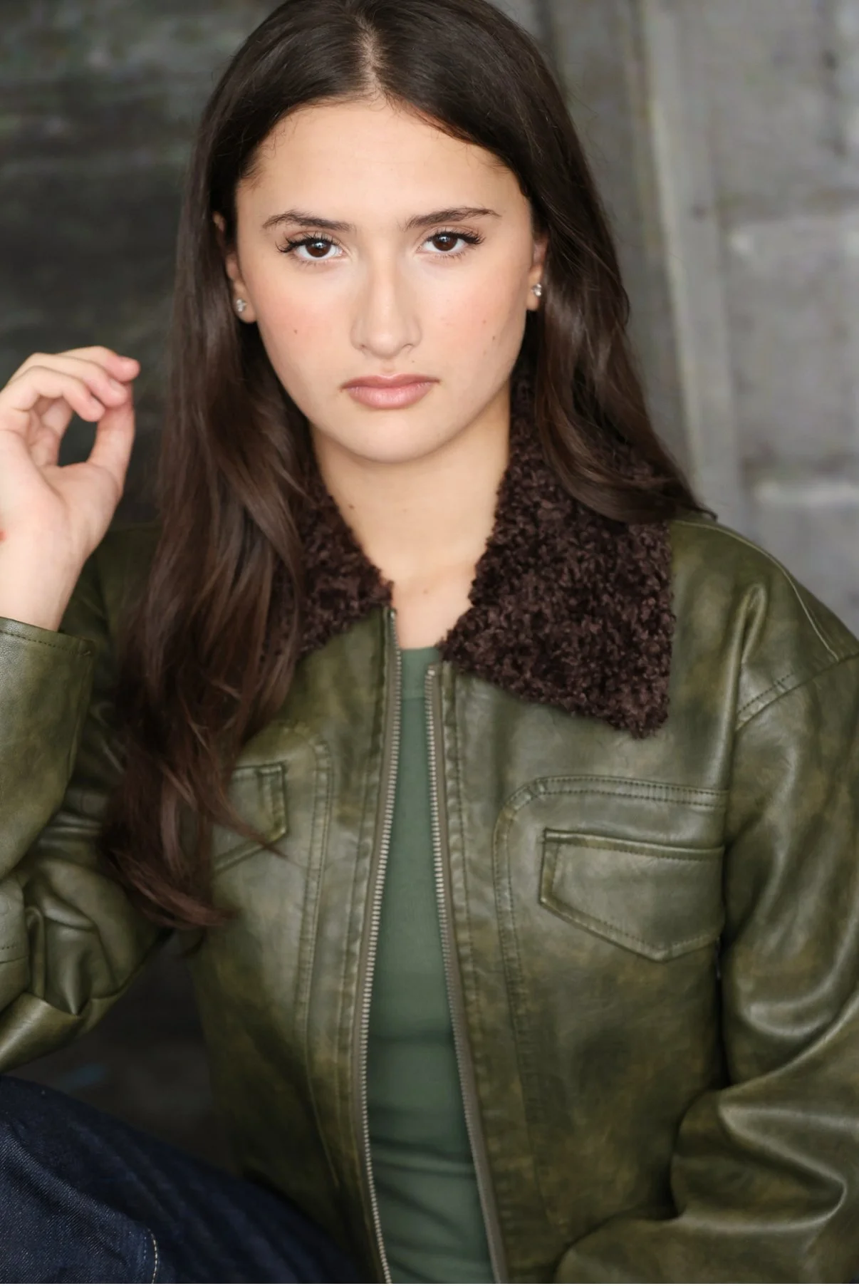 A young woman with long brown hair, wearing a green jacket with a brown sherpa collar, looking directly at the camera with a neutral expression.
