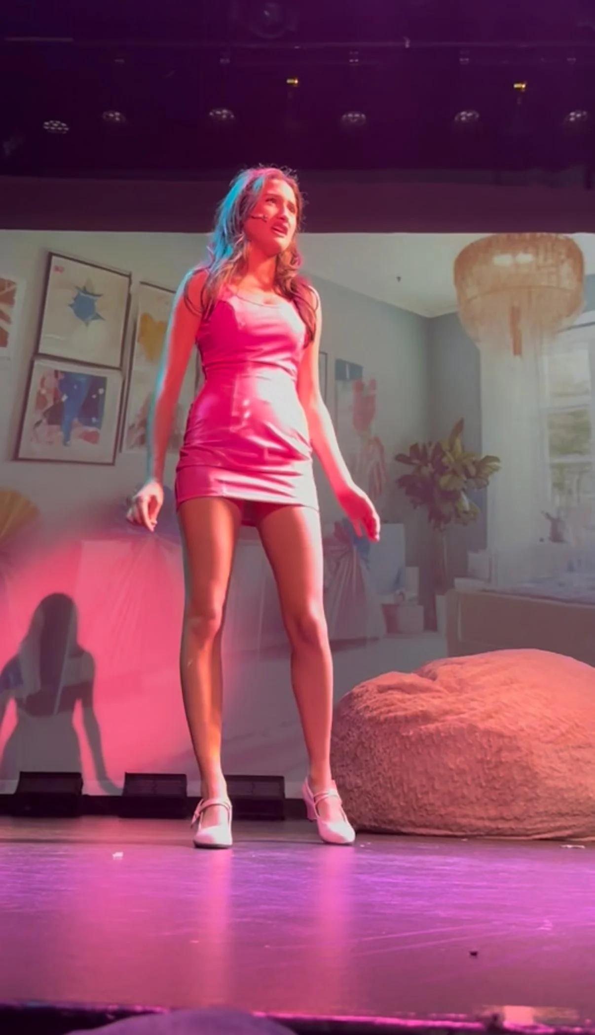 A woman standing on stage, wearing a pink satin dress and high heels, with a pained or emotional expression. The background shows a decorated living room with framed pictures, plants, and a large hanging light fixture.