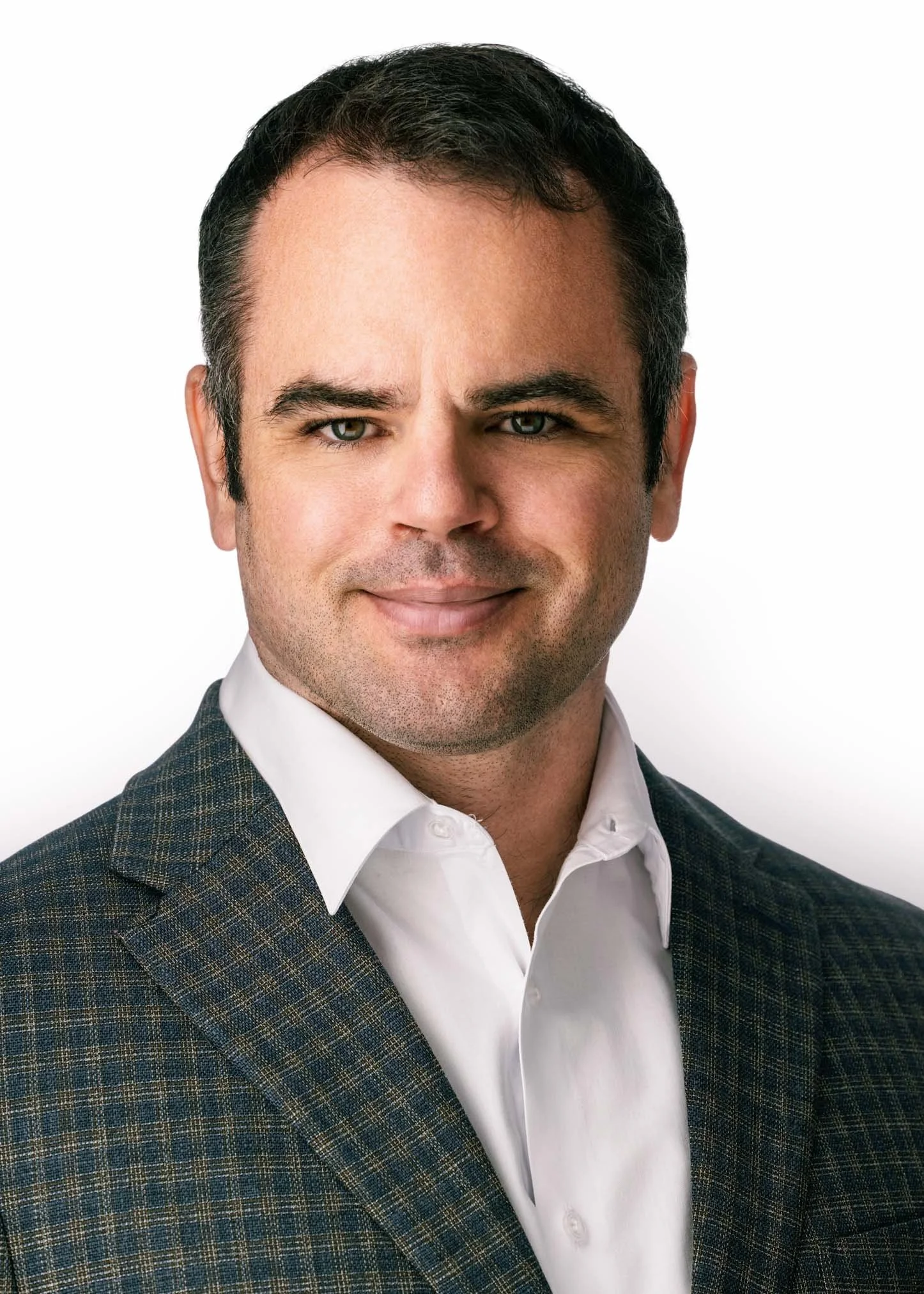 A man with dark hair, light skin, and blue eyes wearing a dark plaid suit jacket and white dress shirt, posed against a white background.