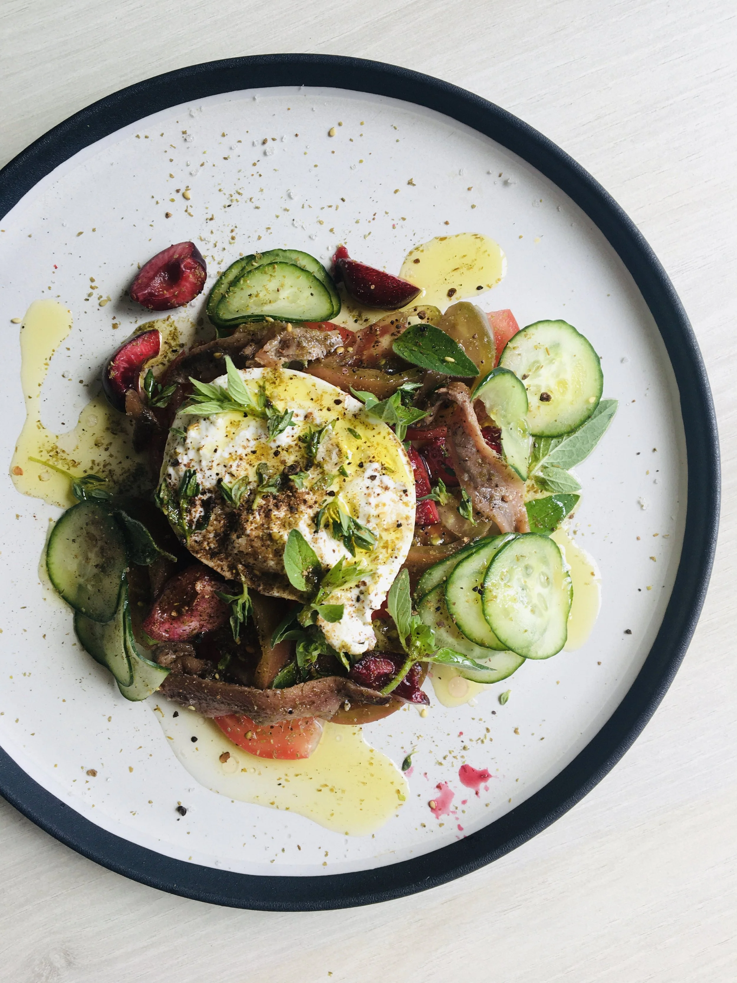 Salade avec œuf mollet, concombre, tomate, herbes, légumes variés, assaisonnement et huile d'olive, présentée dans une assiette blanche avec bord noir.