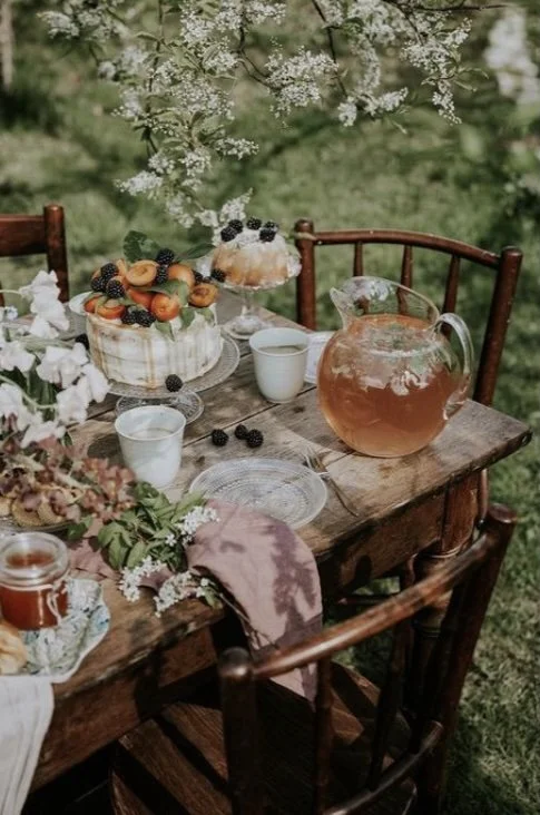 Table rustique en bois avec une carafe de thé rose, des tasses, un gâteau décoré de fruits, des biscuits, et un bouquet de fleurs, dans un jardin avec des fleurs blanches en arrière-plan.