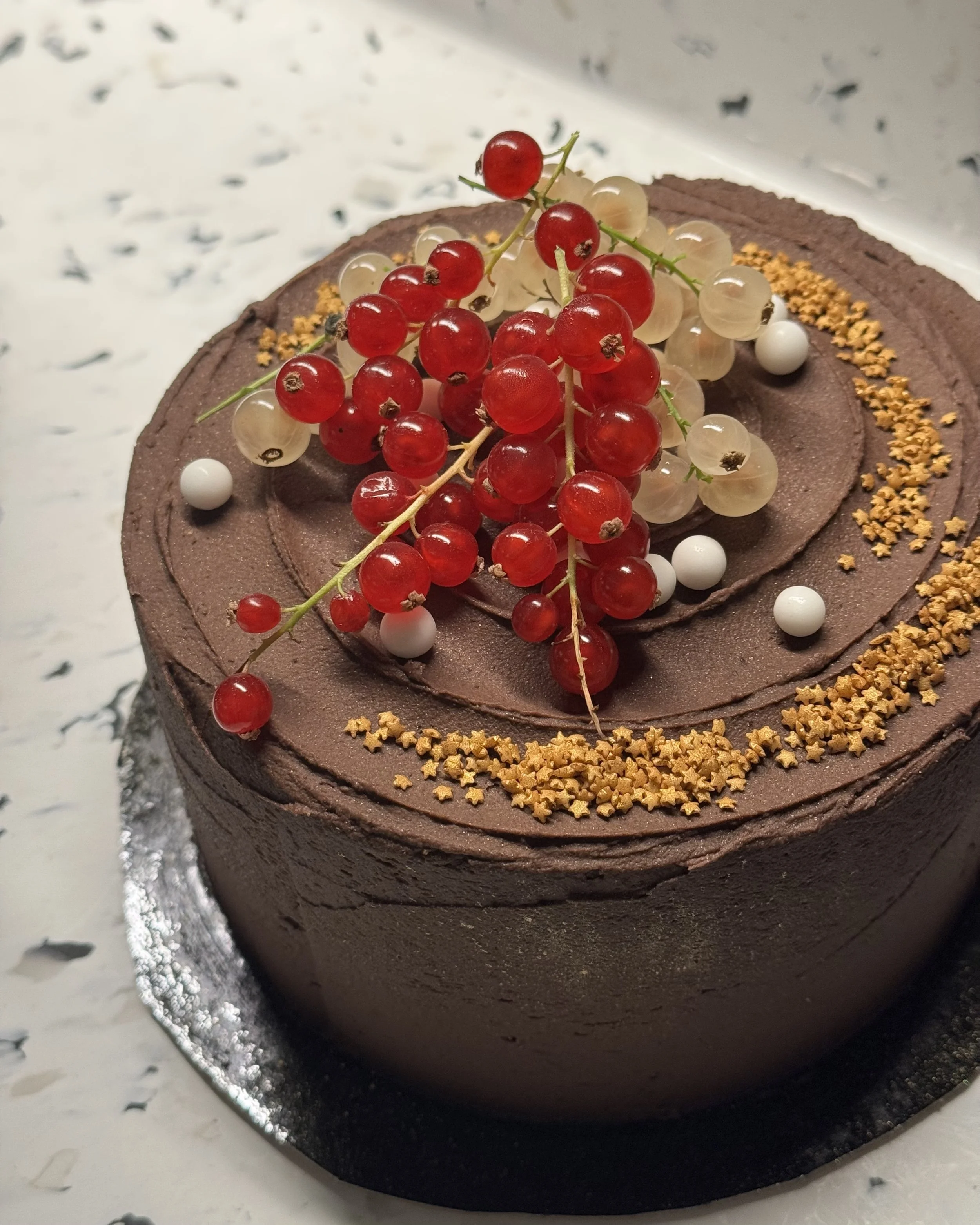Gâteau au chocolat décoré de groseilles rouges, gelées blanches, perles blanches, et petites étoiles en biscuit dorées, sur un socle noir avec un fond blanc marbré.
