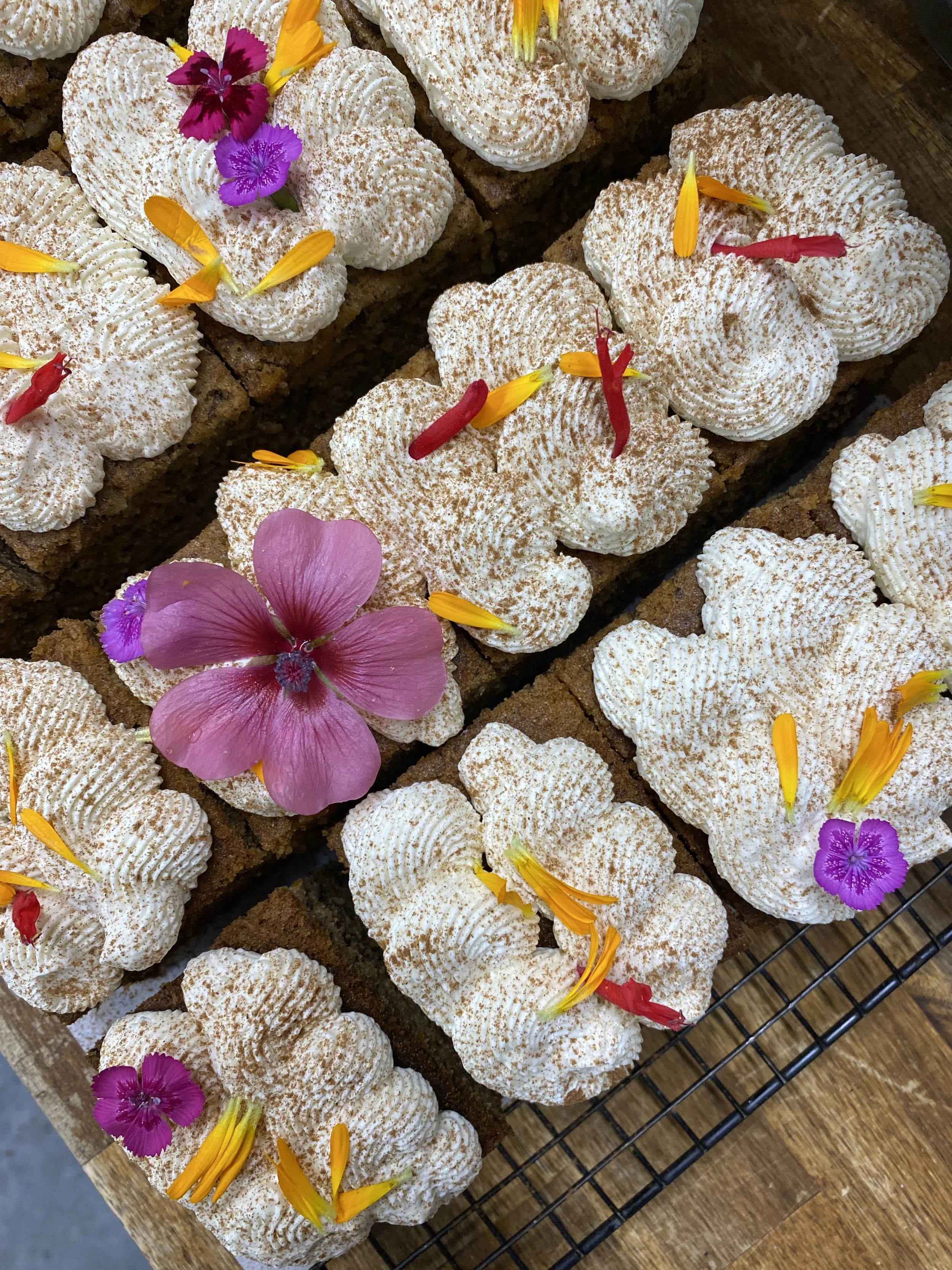 Gâteaux décorés avec de la crème, des fleurs comestibles jaunes, roses, violettes et rouges, sur une grille en bois.