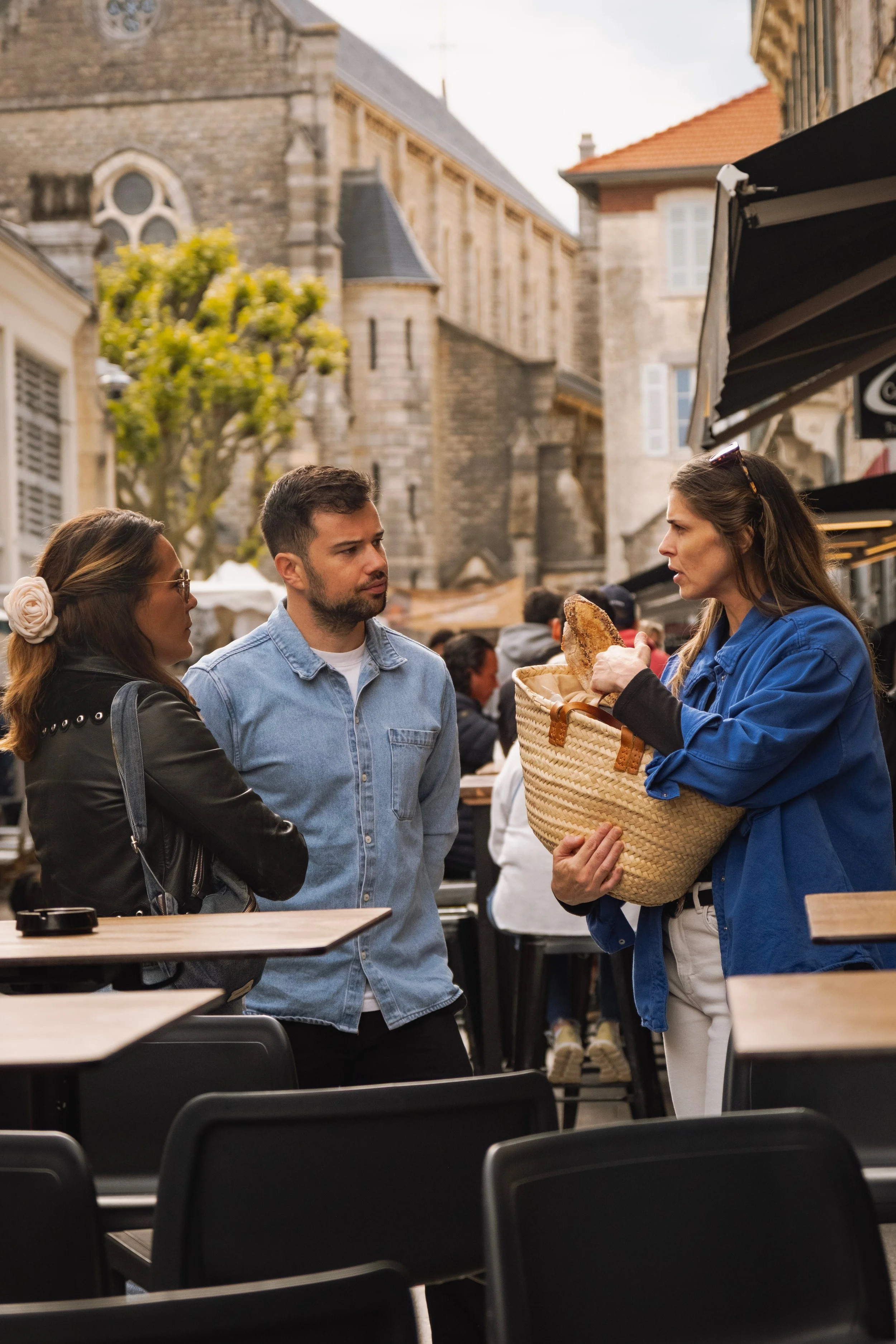 Trois personnes conversent sur une terrasse en ville, avec un bâtiment ancien en pierre en arrière-plan. Une femme avec un panier de pain semble expliquer quelque chose à un homme et une autre femme.