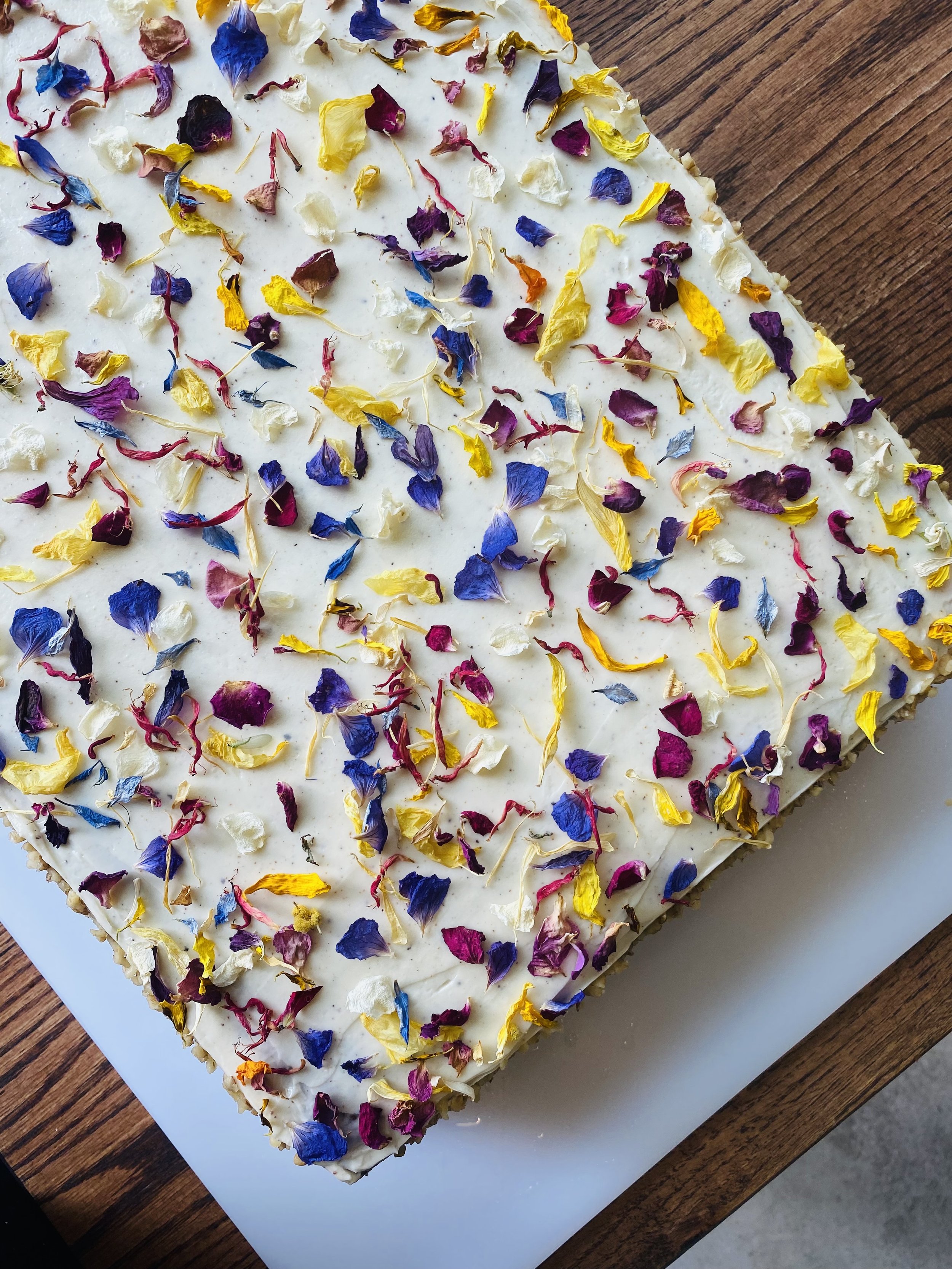 Gâteau décoré avec des pétales de fleurs colorés sur une surface blanche, placé sur une table en bois.