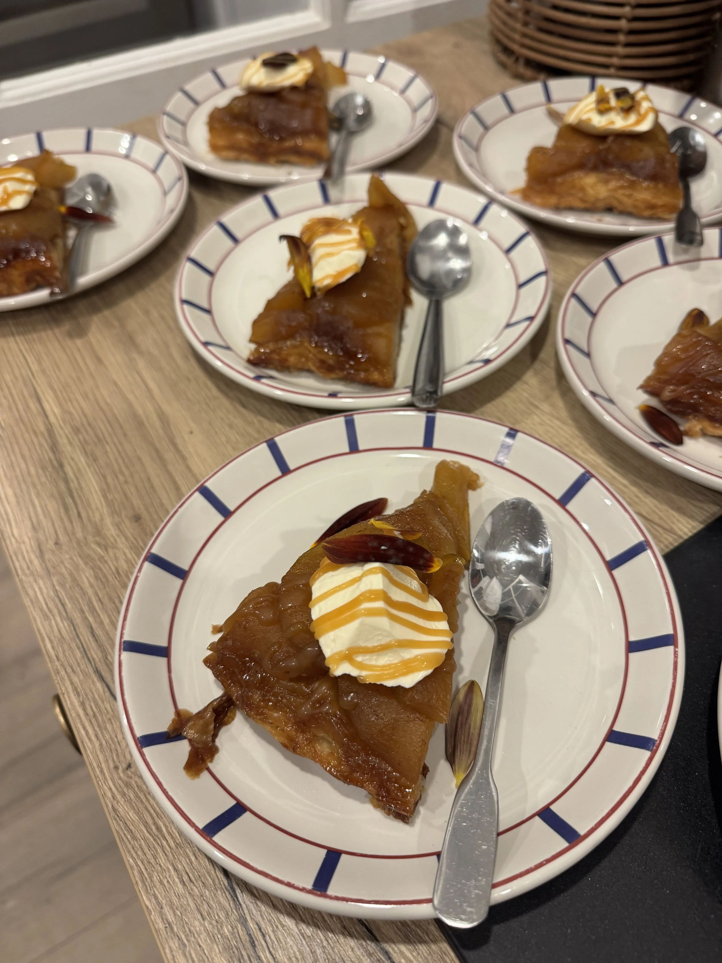 Une part de tarte aux pommes garnie de crème, sauce caramel et quelques amandes, servie dans une assiette blanche avec un bord à rayures rouges et bleues, accompagnée d'une petite cuillère en métal, sur une table en bois.