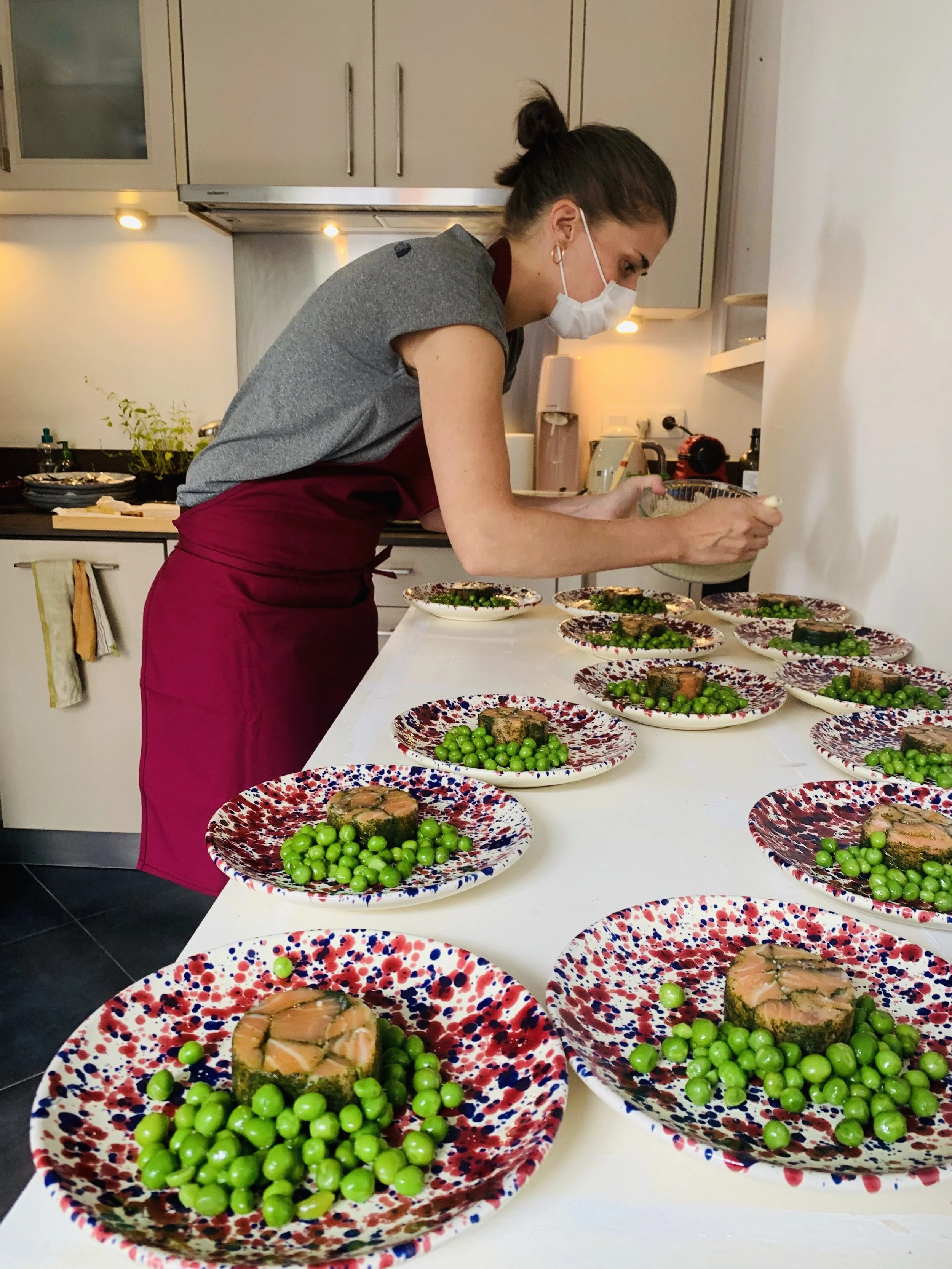 Une femme portant un masque, une robe grise et un tablier rouge, prépare des plats dans une cuisine moderne, avec plusieurs assiettes colorées contenant de la nourriture verte sur une table