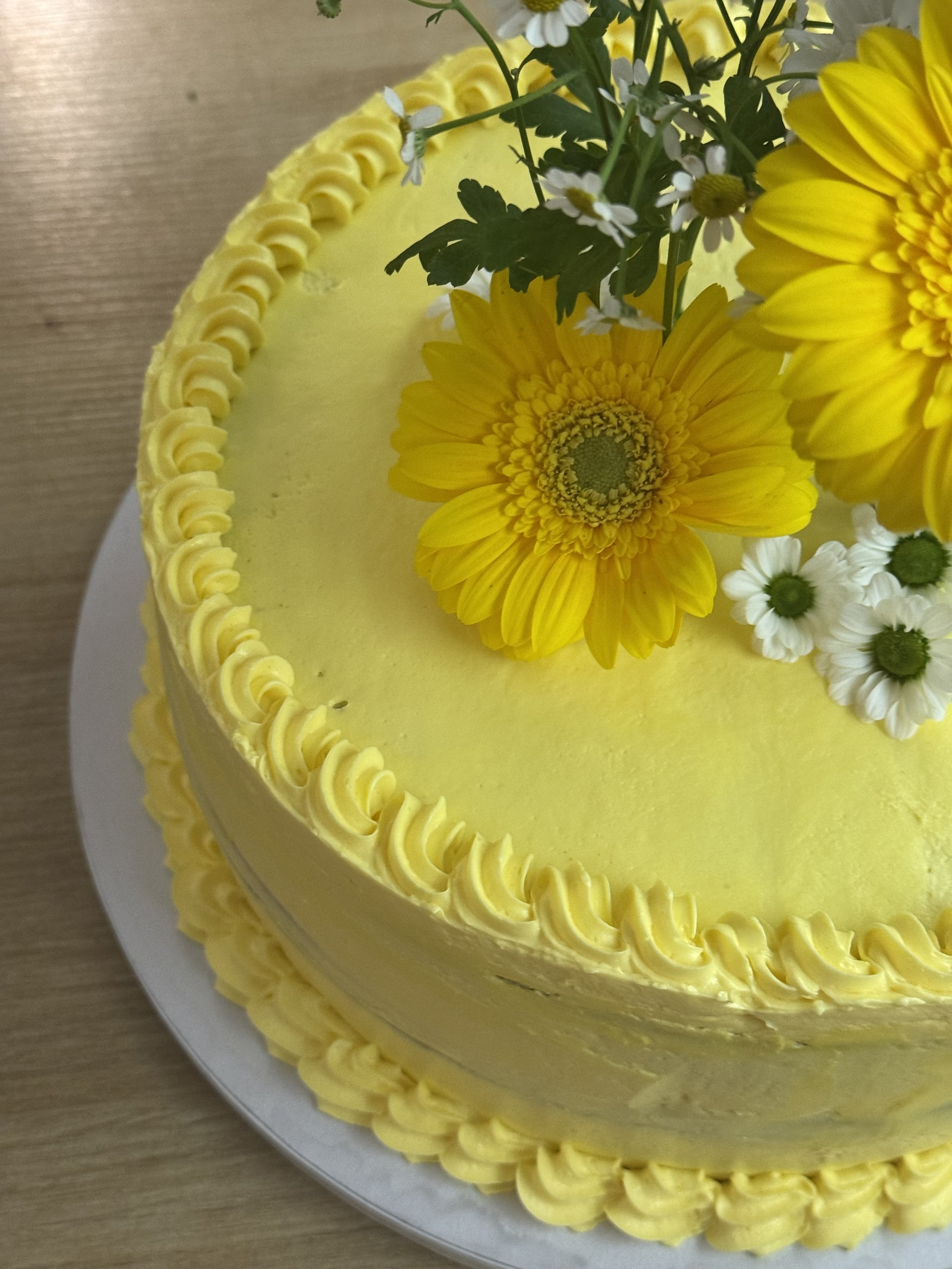Gâteau jaune décoré de fleurs, dont des marguerites, avec un bord en décoration de crème jaune.