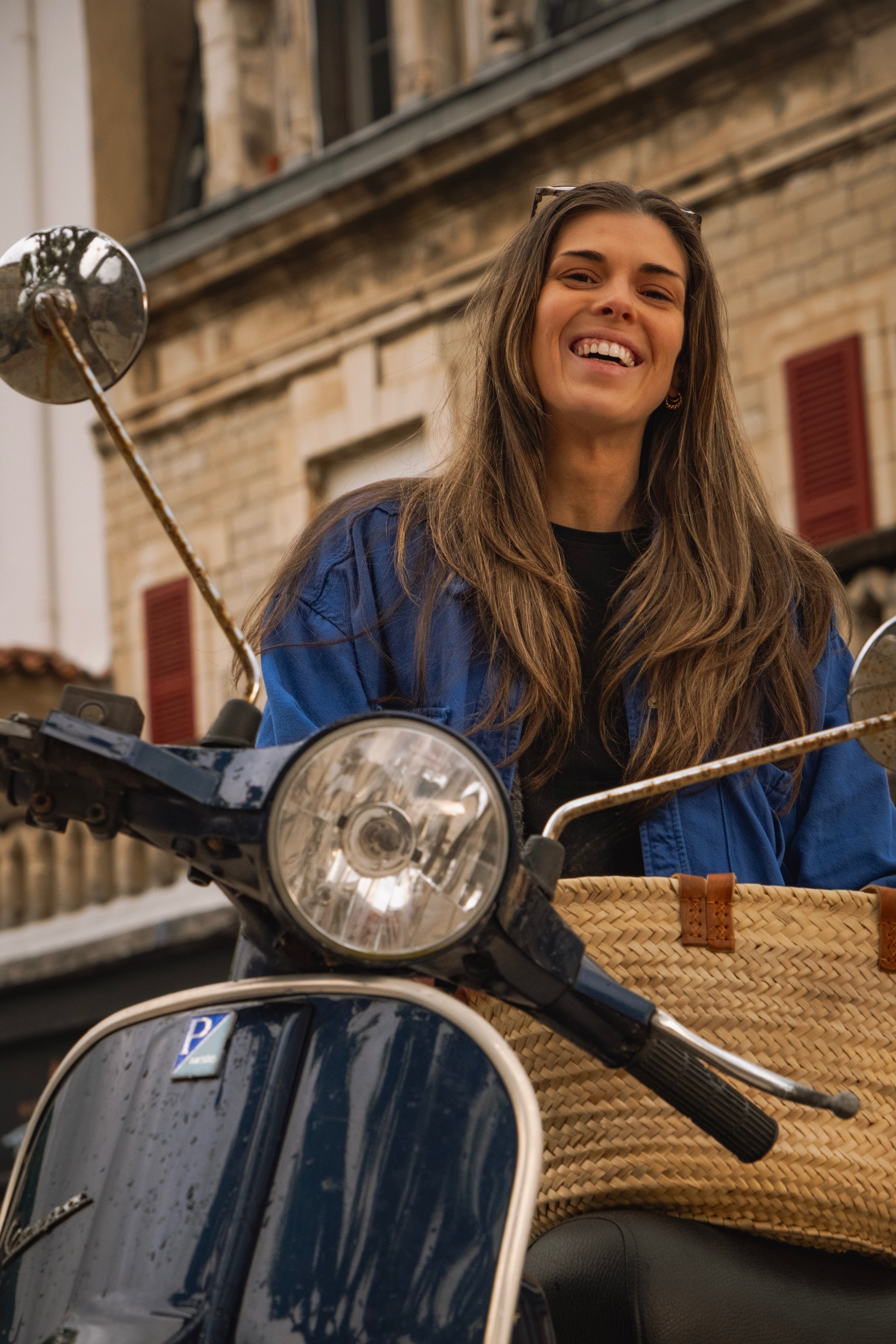 Une femme souriante portant une veste bleue, assise sur une moto, avec un bâtiment en pierre en arrière-plan.