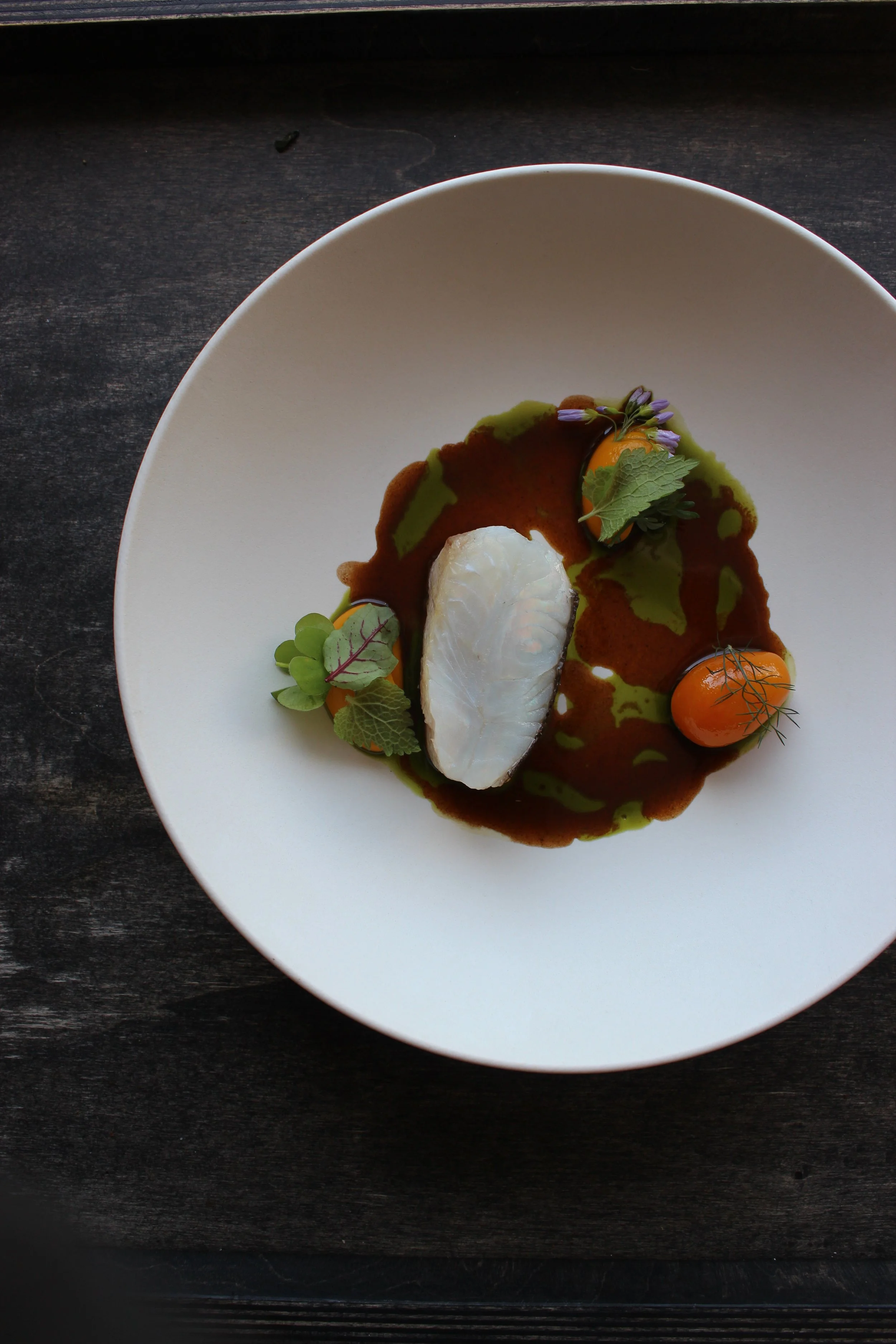 Assiette blanche contenant un plat gastronomique avec un œuf poché, des petits légumes, des herbes, des sauces vertes et brunes, présentation élégante sur une surface sombre.
