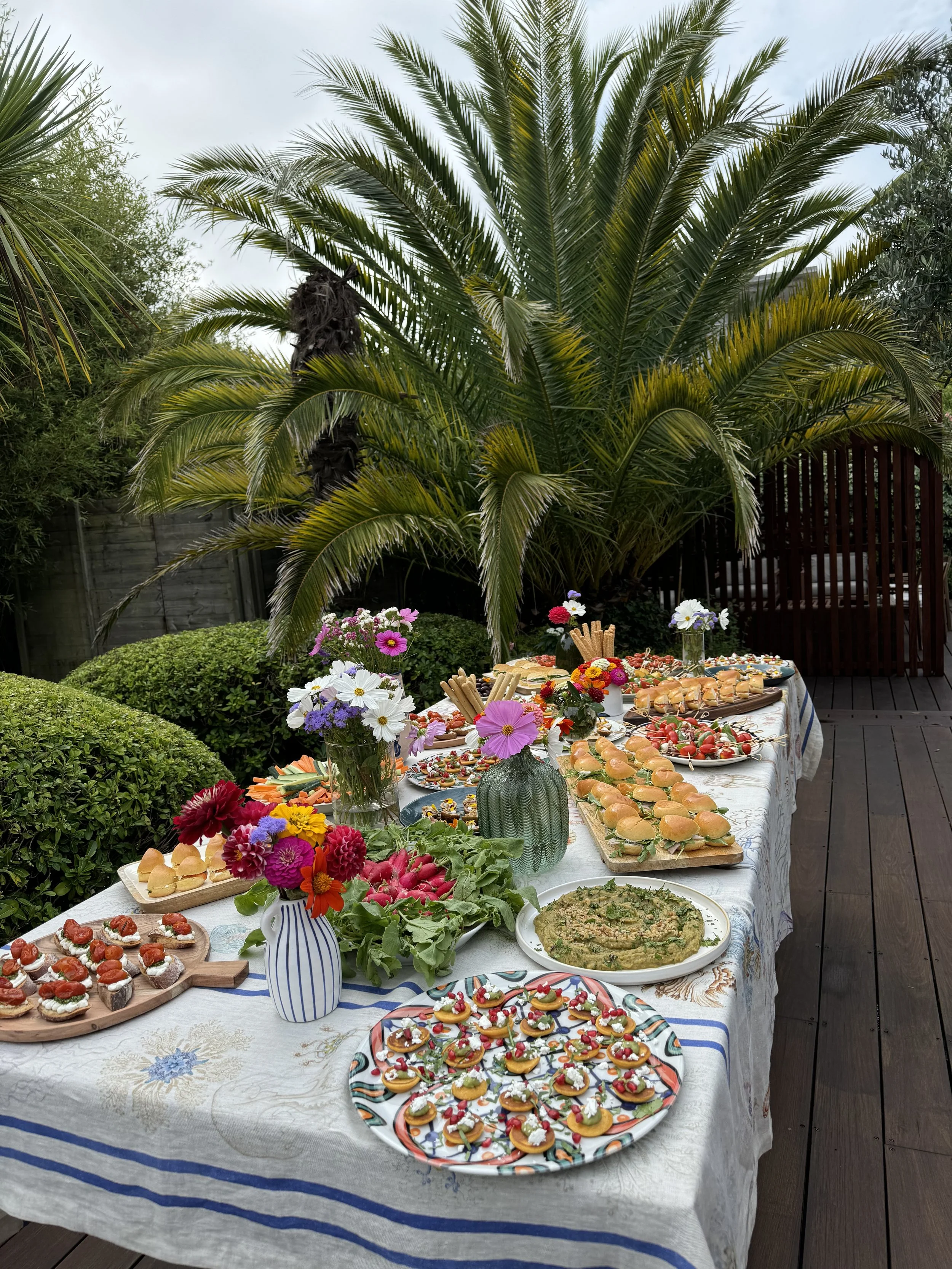Un buffet de fête avec diverses amuse-gueules et desserts colorés, décoré avec des fleurs, située dans un jardin avec des palmiers.