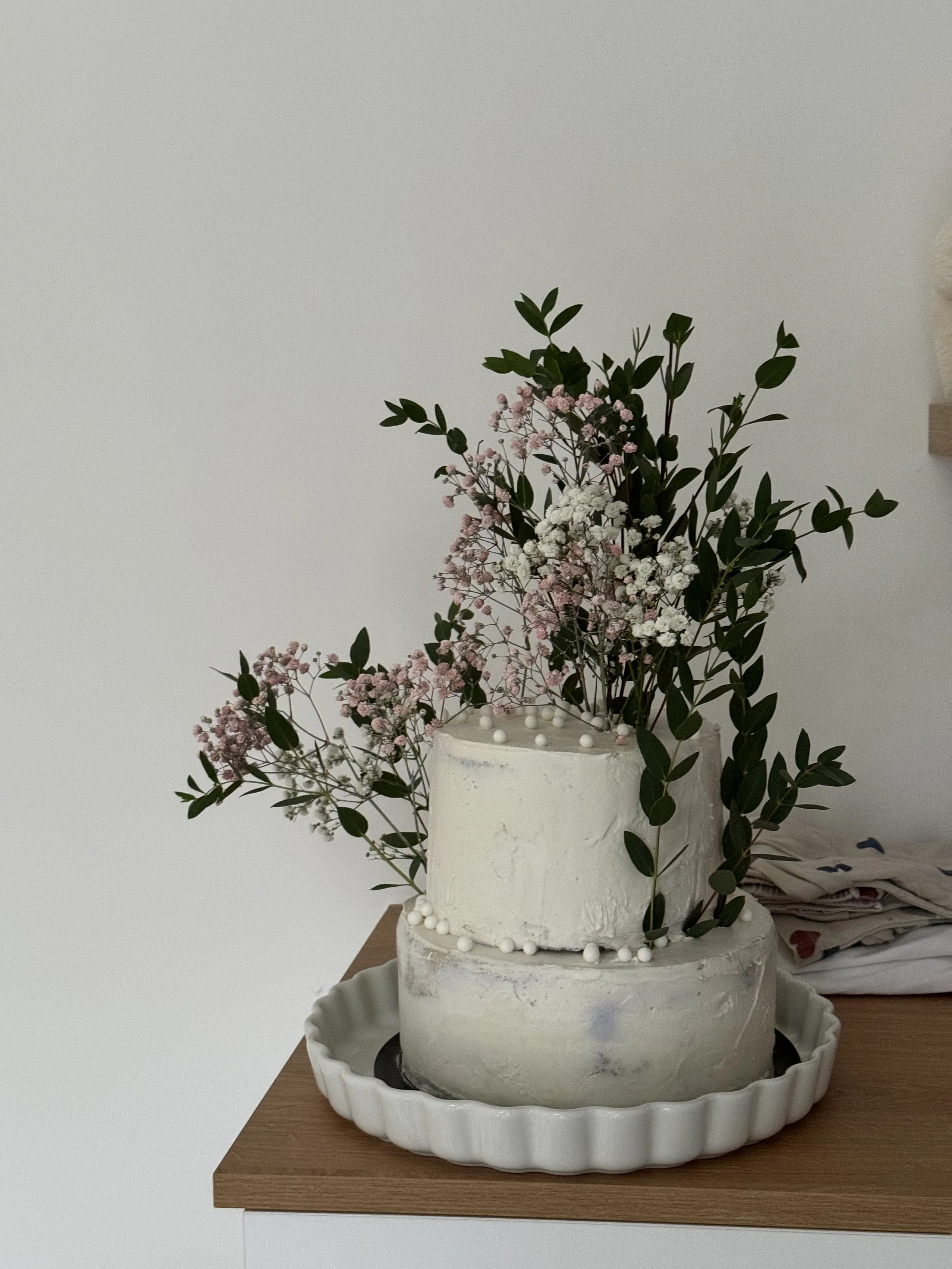 Gâteau blanc décoré de fleurs et de feuilles vertes, placé dans un plat blanc, sur une table en bois.