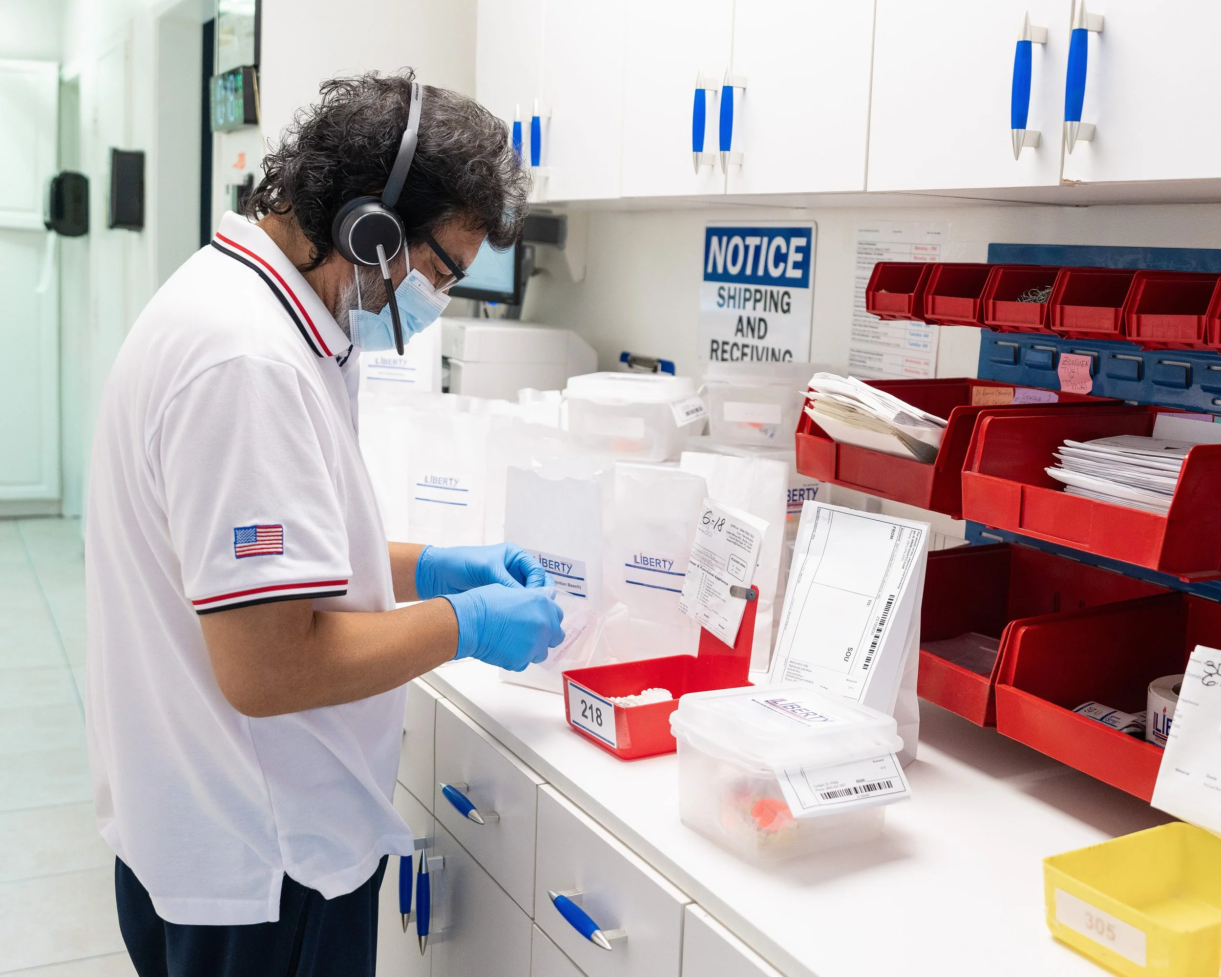 A healthcare worker wearing a white Uniform with an American flag patch on the sleeve, blue gloves, glasses, a mask, and a headset, standing in a medical facility and managing shipping labels or packages at a counter with red and white bins containing documents and supplies.