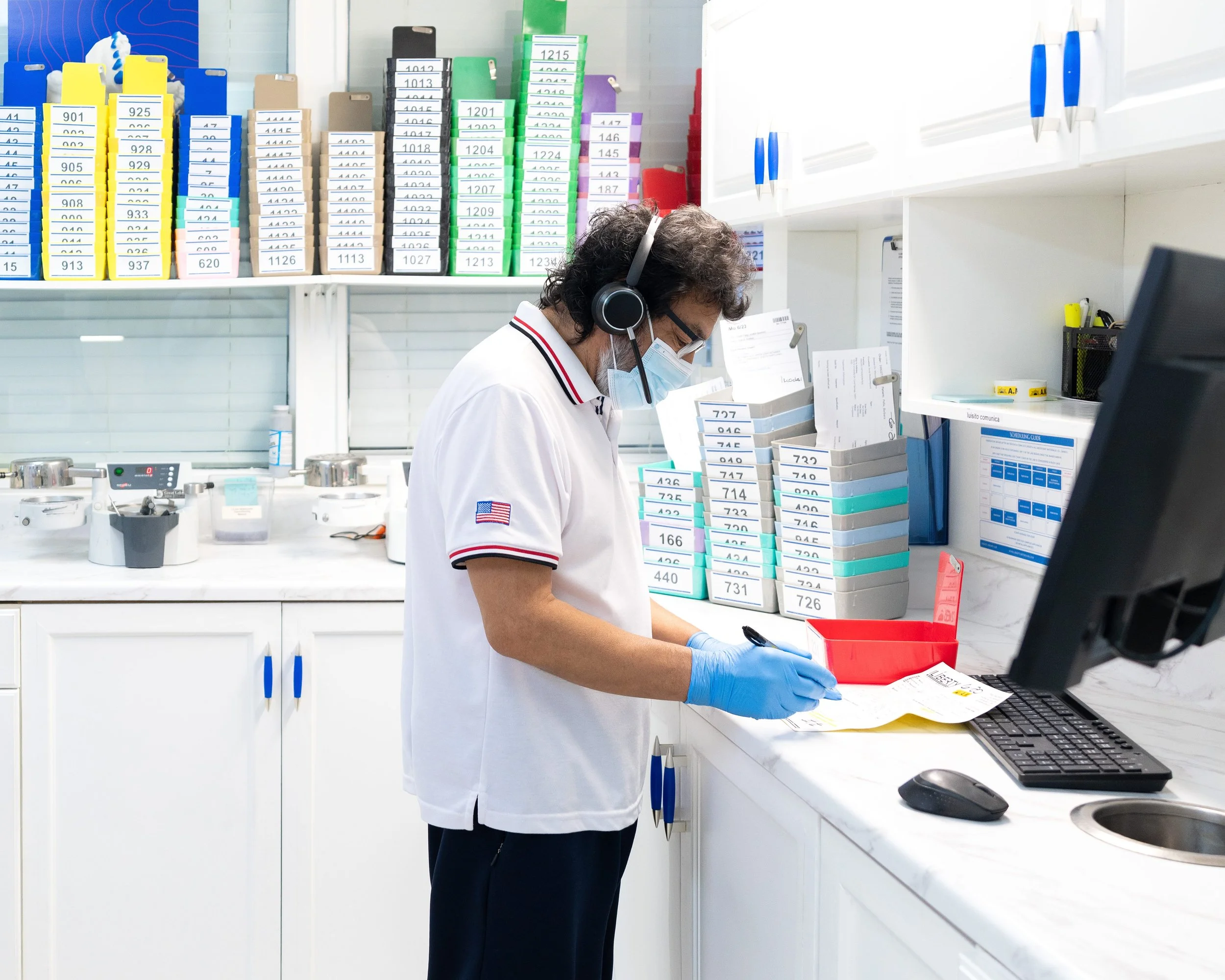 A man wearing a white shirt, face mask, glasses, and blue gloves working at a pharmacy or medical lab. He is standing at a counter with organized boxes and paperwork, with a background of shelves filled with colored file folders and office supplies.