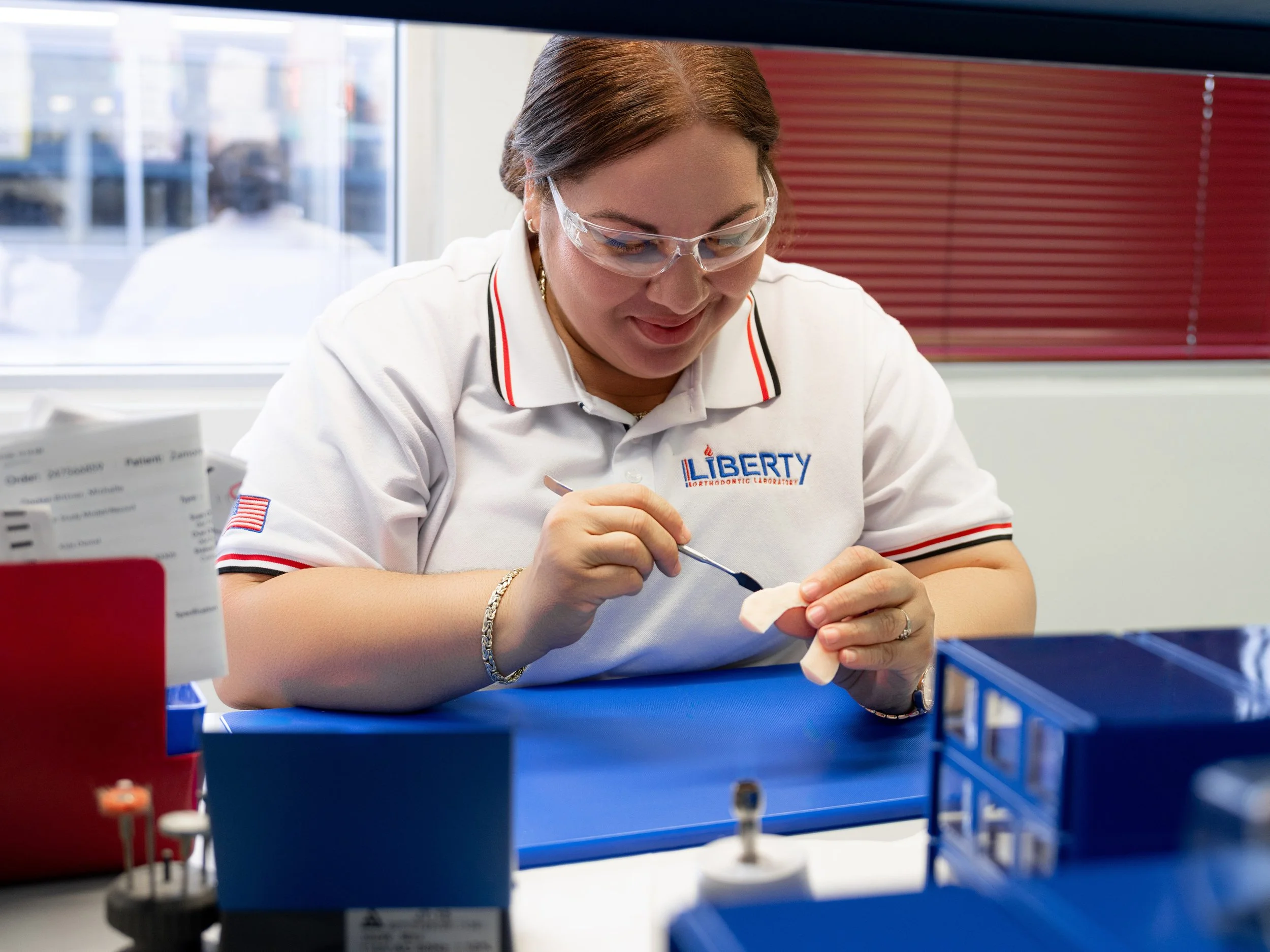 A woman with brown hair wearing glasses and a white shirt with 'Liberty Orthodontic Laboratory' embroidered on it, working on a dental appliance at a laboratory station.