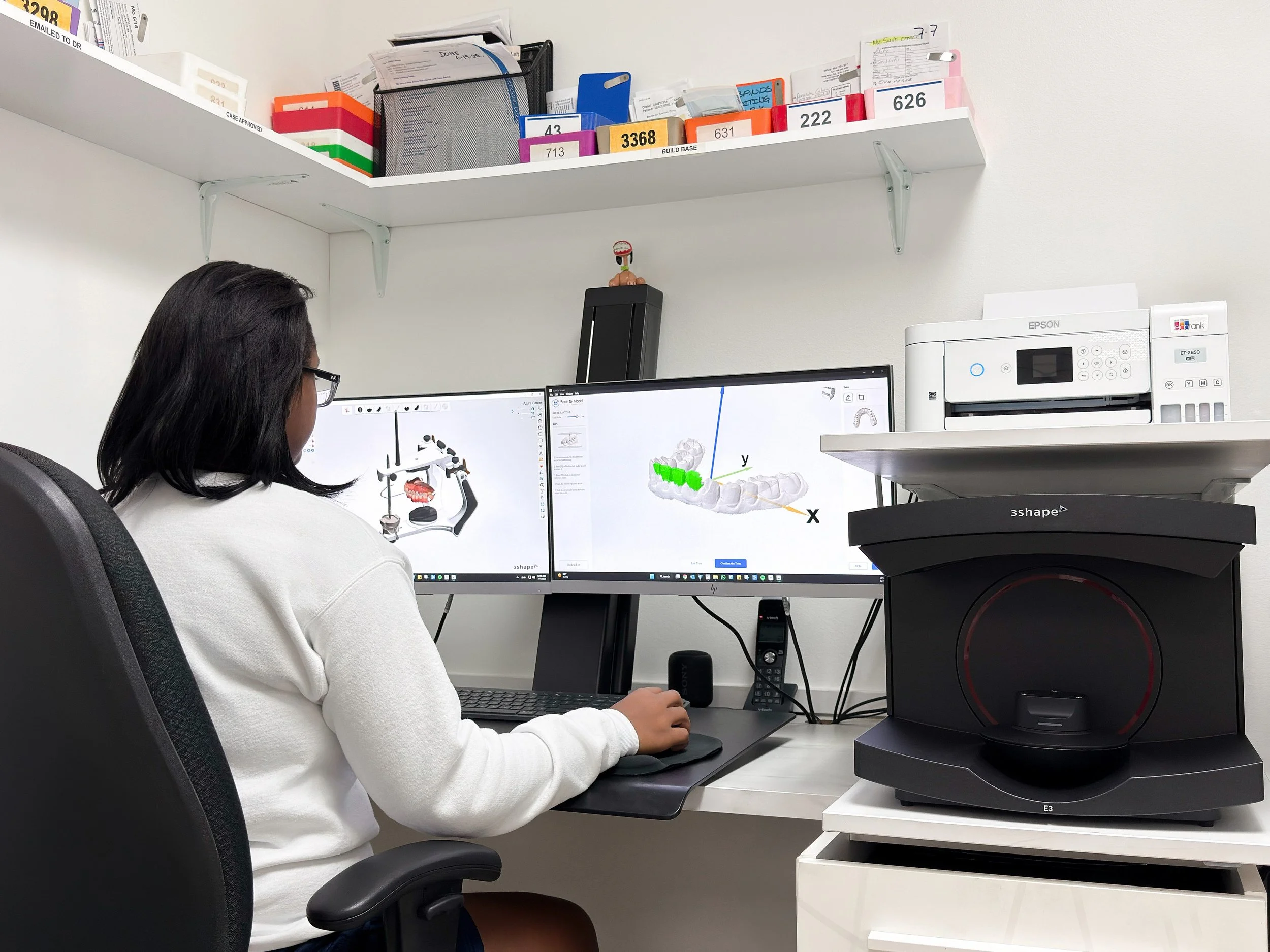 A woman with shoulder-length black hair and glasses working at a desk with dual computer monitors displaying a 3D model of teeth and gums. The desk also has a black speaker, a filing cabinet, and a white shelf with labeled folders and office supplies in the background.