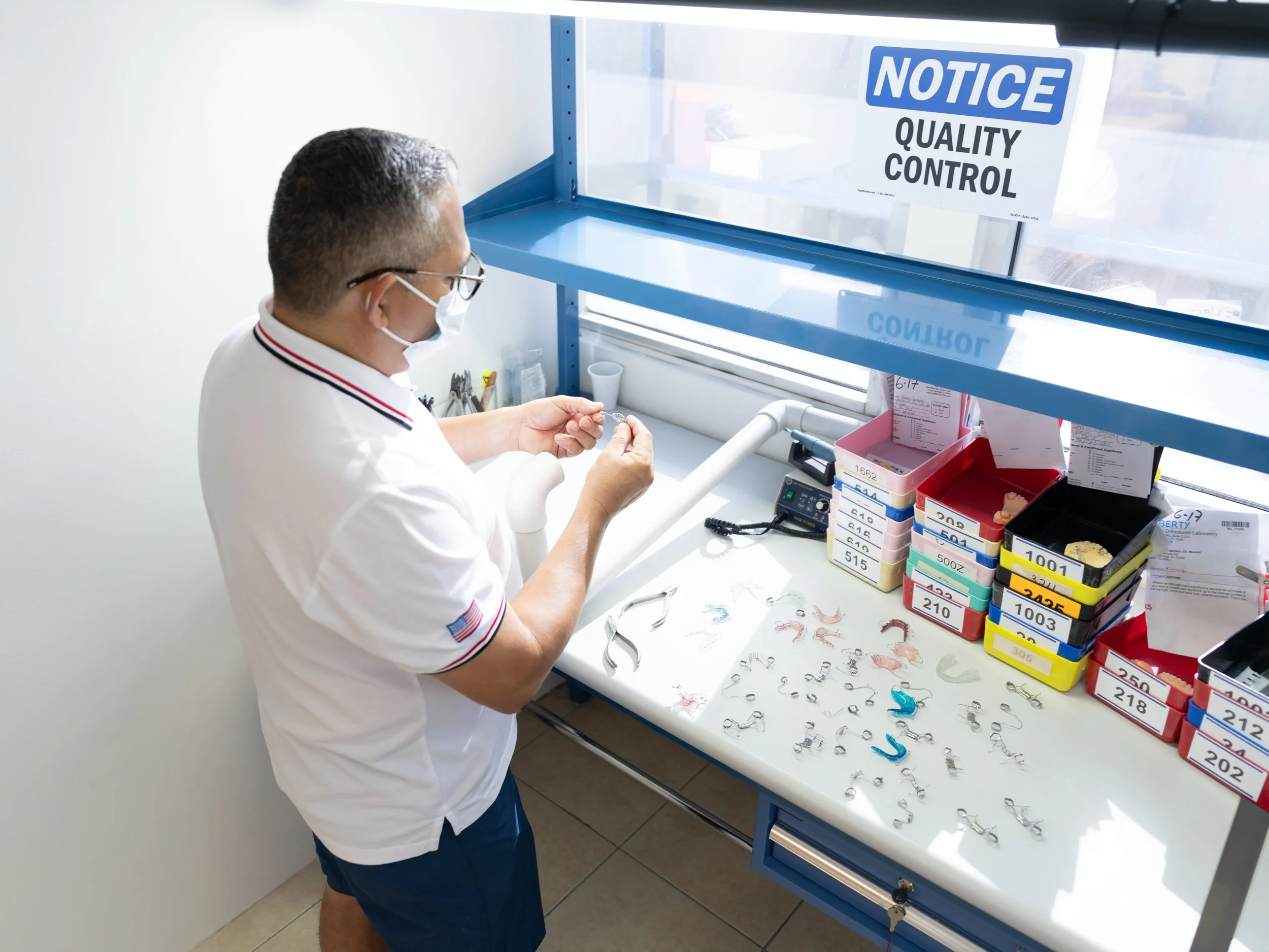 A man wearing glasses, a face mask, a white polo shirt, and blue shorts is examining dental or orthodontic appliances on a white workbench with various labeled drawers containing small parts and tools. There is a blue shelf with a sign that reads 'NOTICE QUALITY CONTROL' in the background.