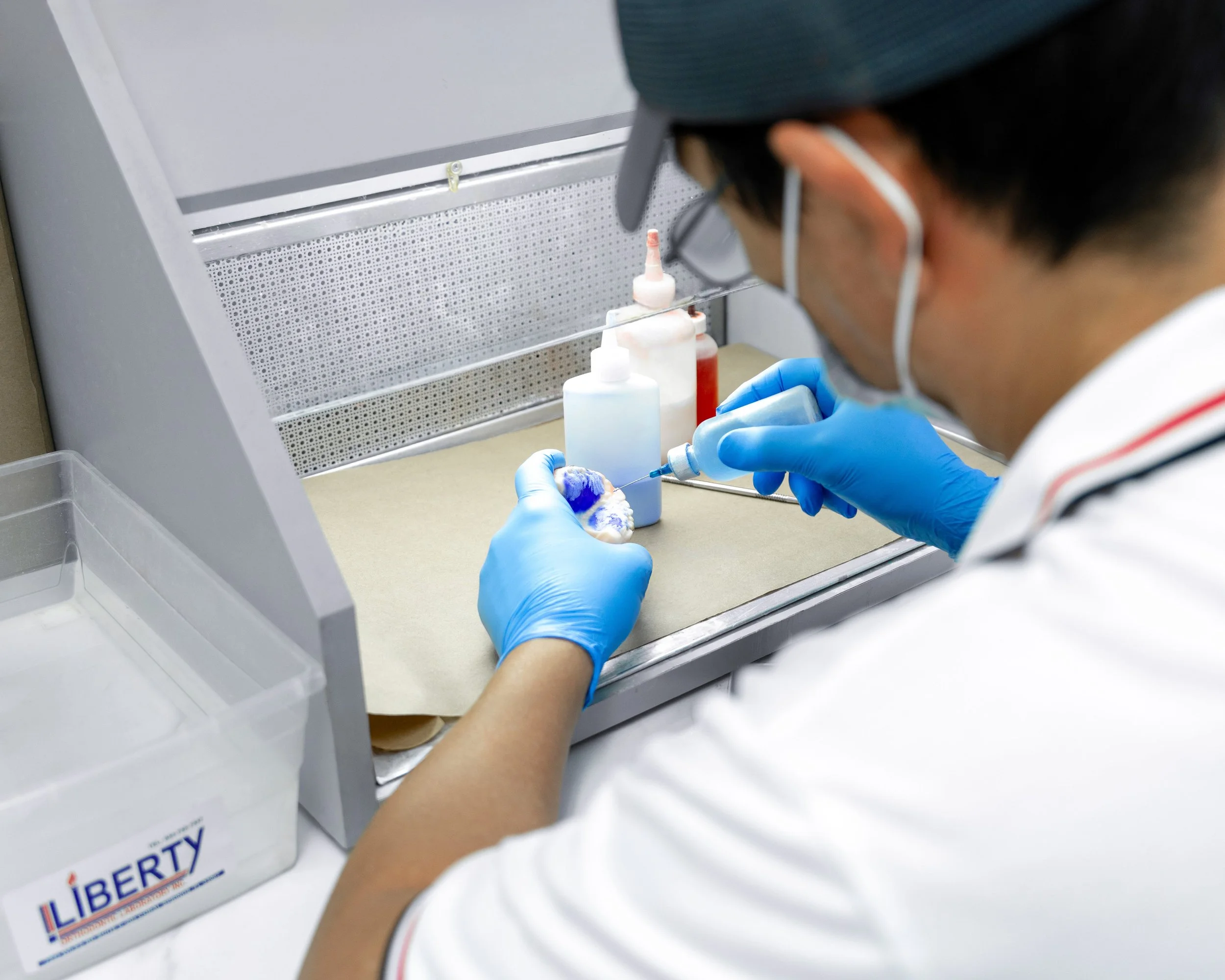 Person wearing gloves and a mask inserting a needle into a vial in a clinical setting.