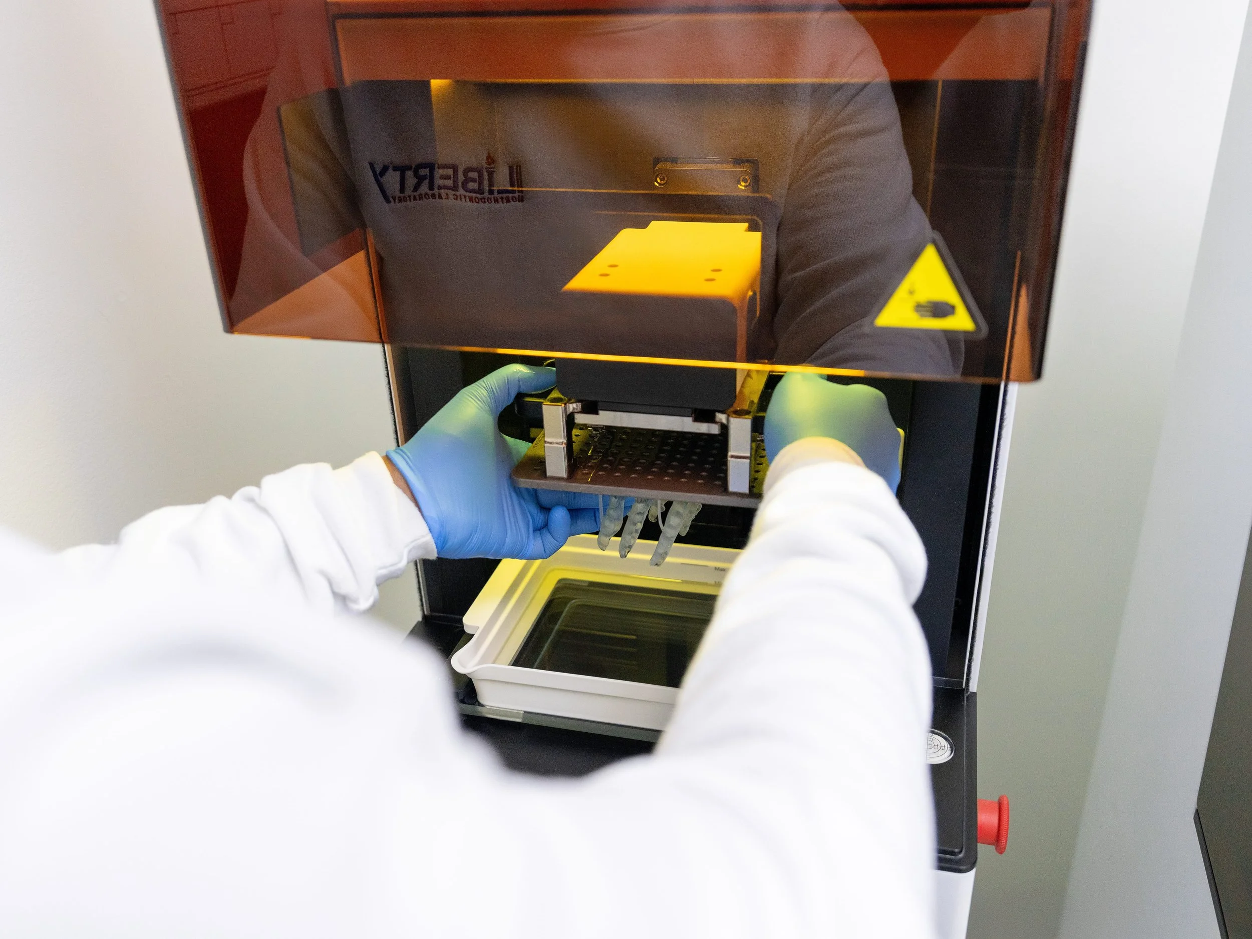 Person wearing blue gloves operating a laboratory instrument with an orange cover, possibly a 3D printer or a scientific device, in a lab setting.