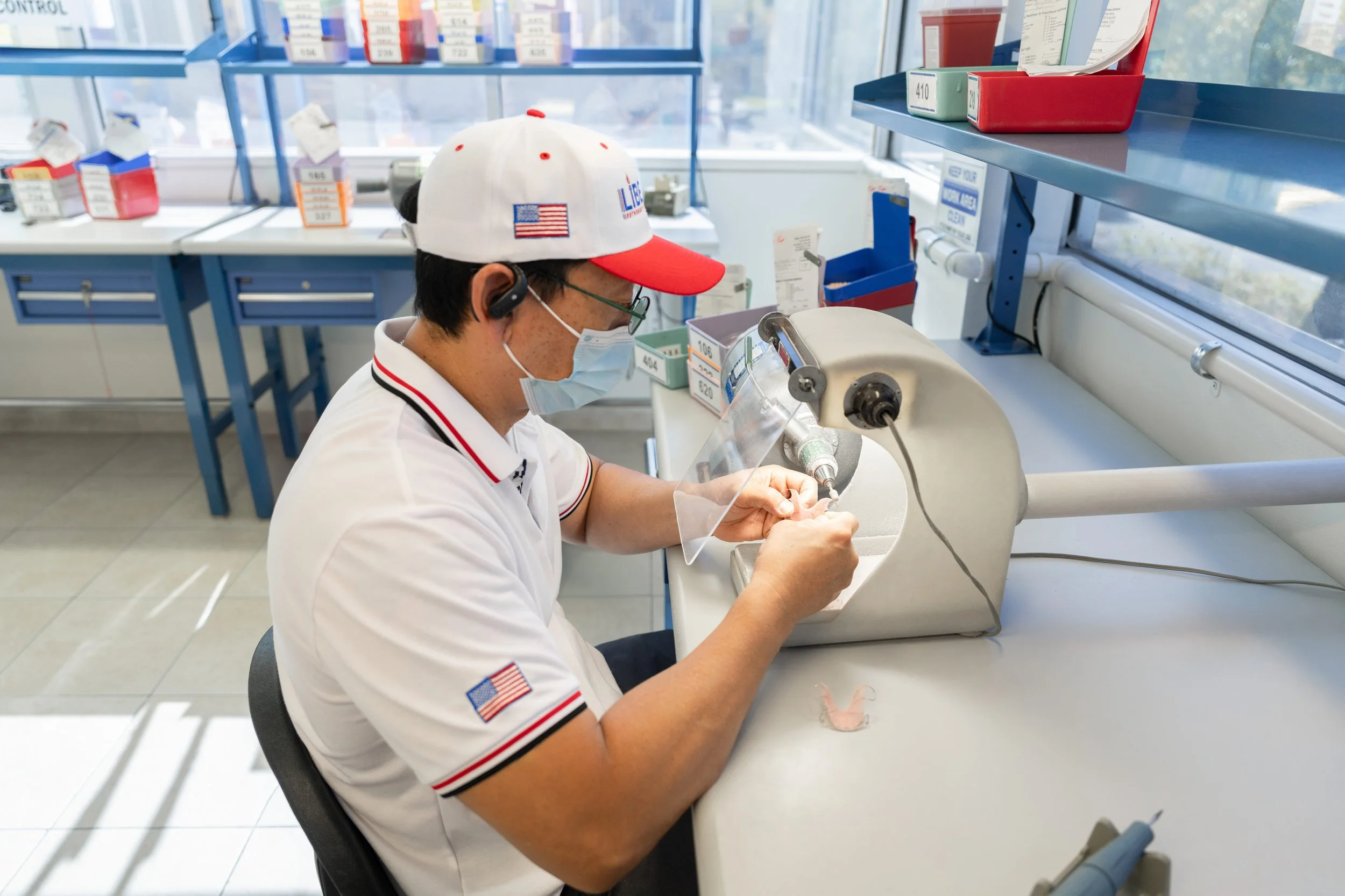 A person wearing a baseball cap, face mask, and cellphone earpiece working with medical or laboratory equipment at a white lab bench.