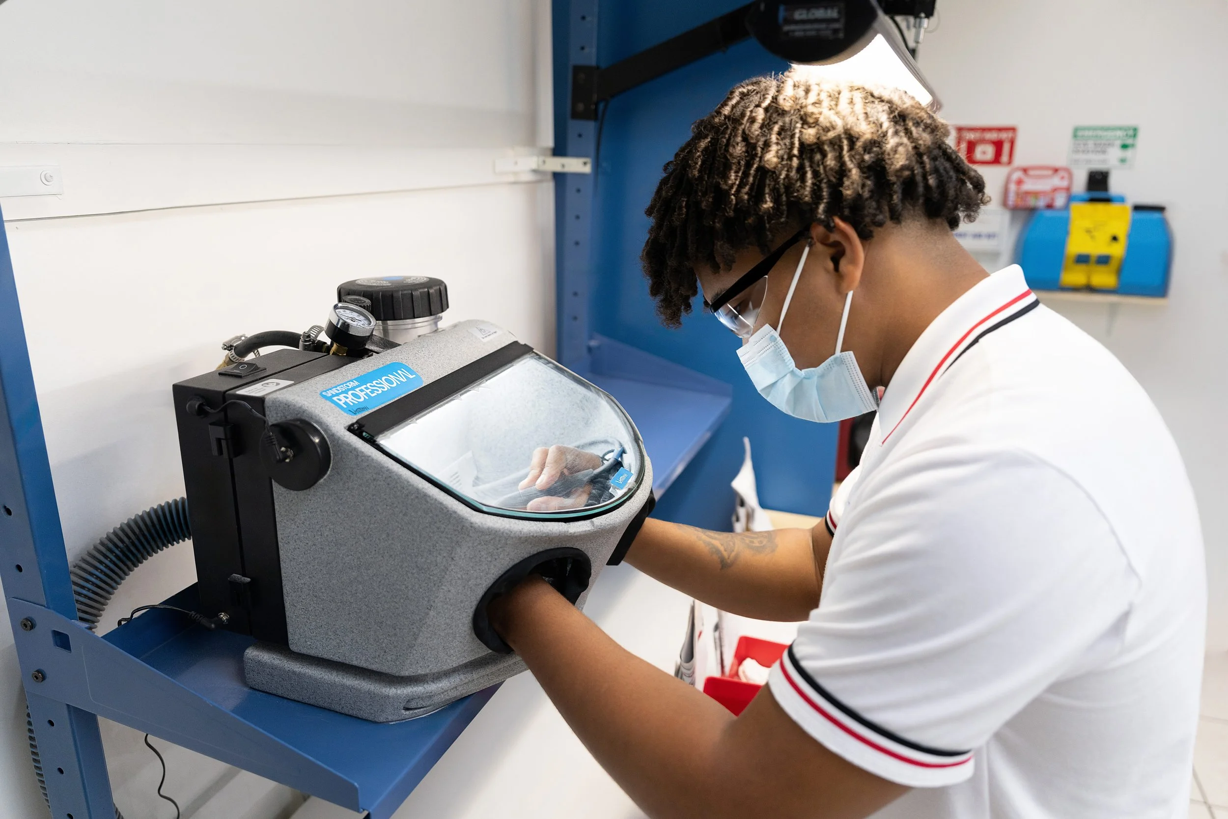 A person with curly hair, glasses, and a face mask working with a piece of equipment in a laboratory.