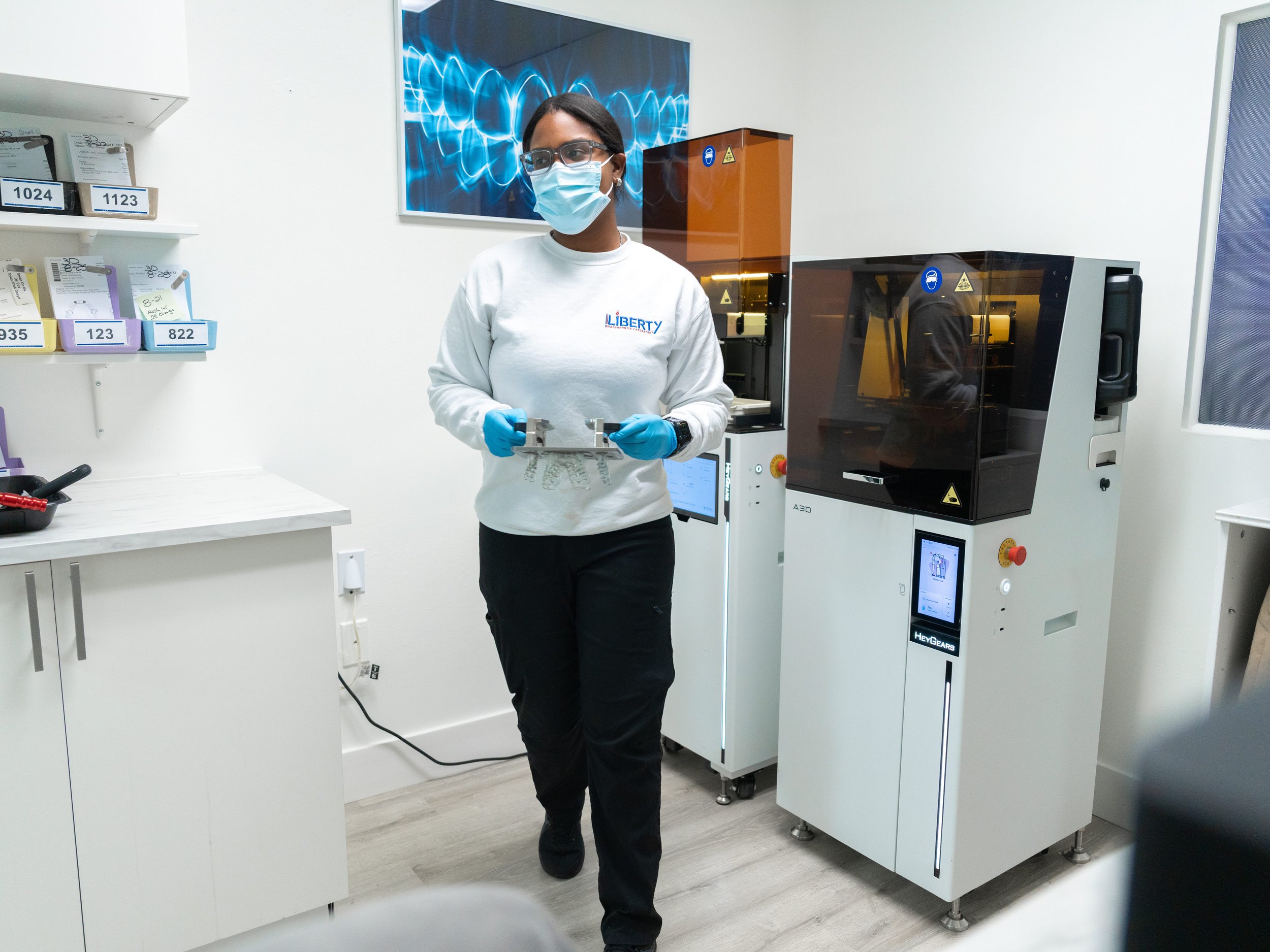 A woman wearing glasses, a face mask, and blue gloves stands in front of laboratory equipment in a medical or research setting. She is holding some objects and is dressed in a white sweatshirt with a logo that reads 'LIBERTY'.
