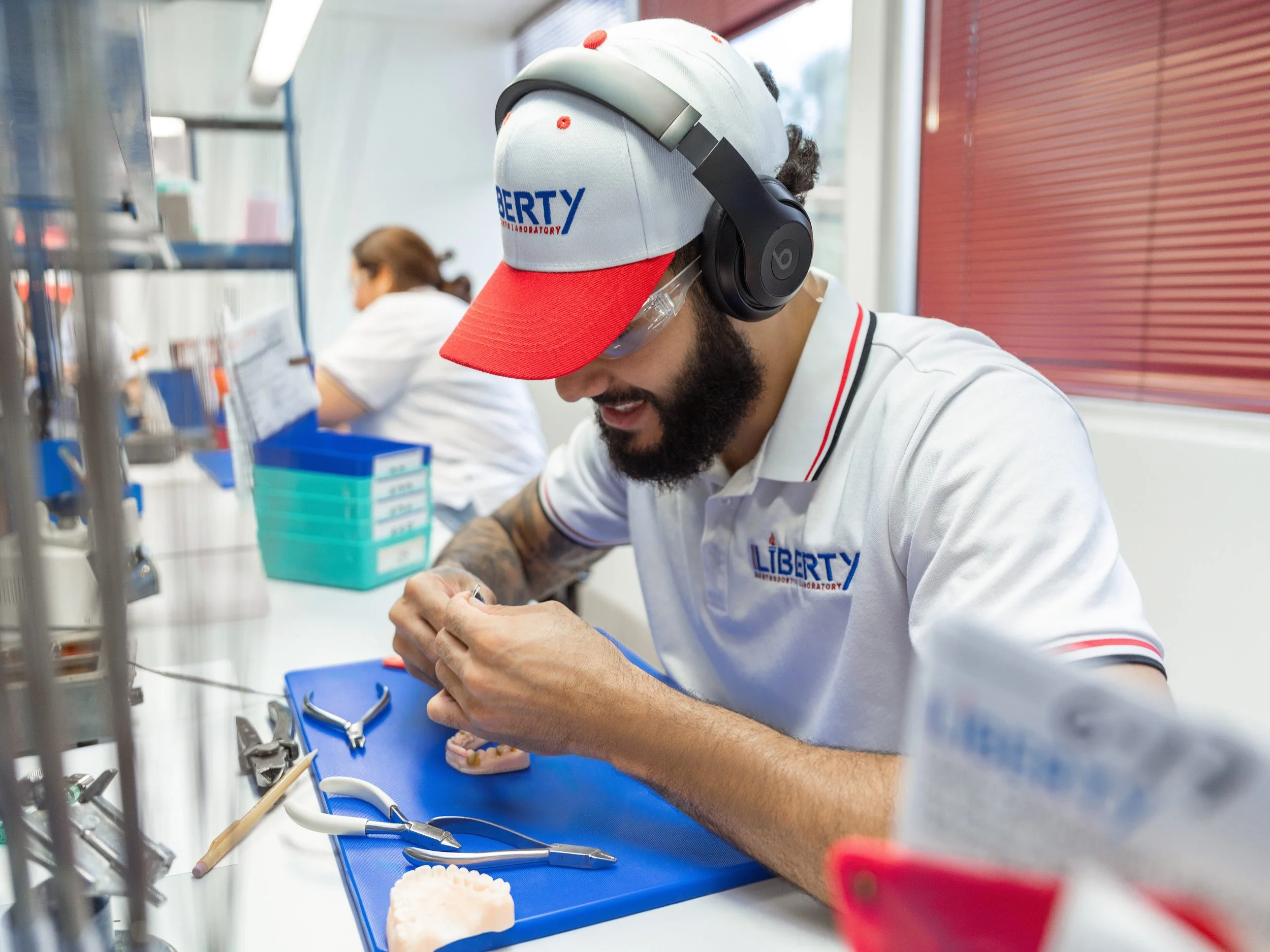 A man wearing a Liberty Laboratory cap and headphones is working on a project at a laboratory bench, surrounded by tools and equipment.