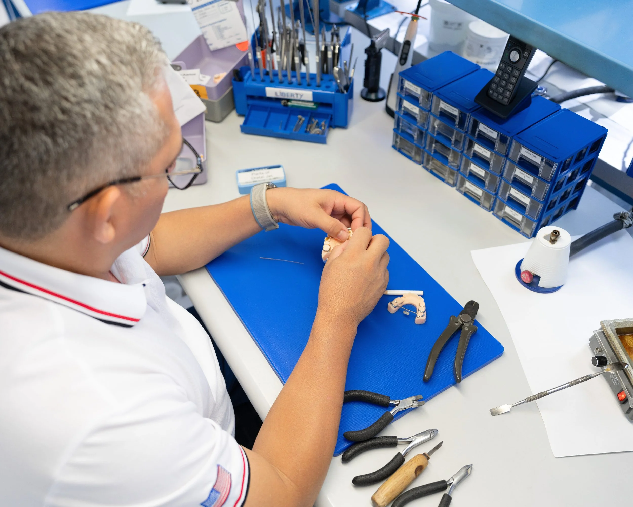 A dental technician working at a workstation with dental tools, a model of teeth, and organizing storage containers.