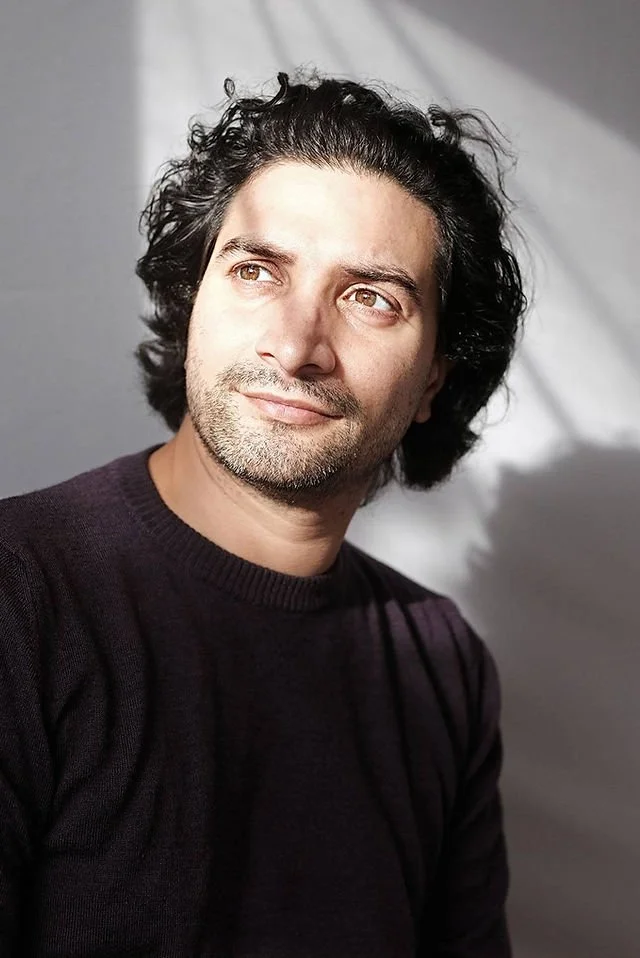 Man with curly dark hair and light skin wearing a black sweater, looking to the side with sunlight on his face, standing in front of a plain background.