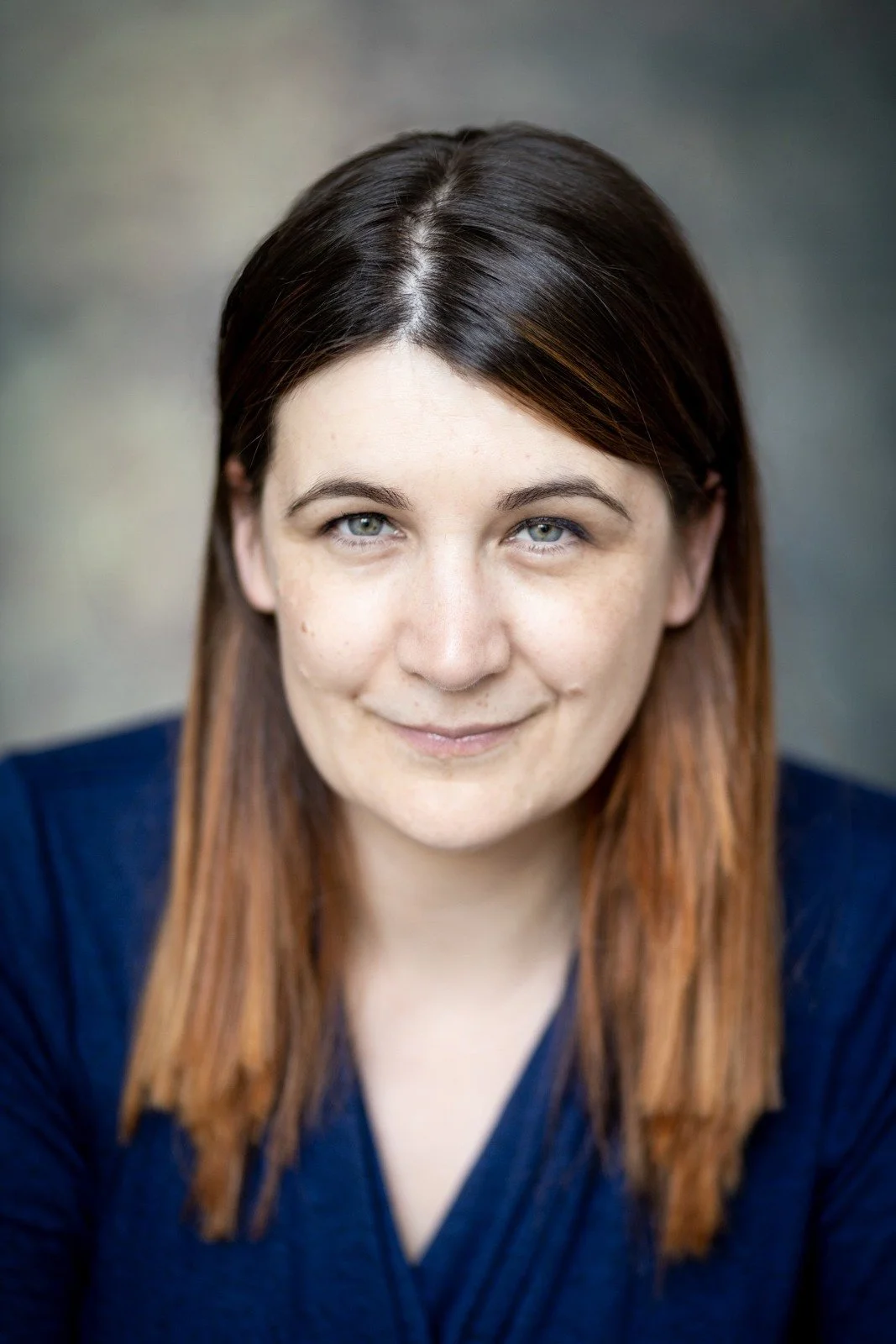 A woman with shoulder-length brown hair with light highlights, wearing a dark blue top, smiling softly at the camera against a blurred neutral background.