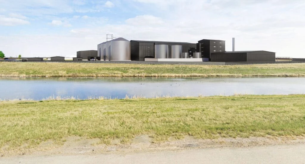 An industrial facility across a body of water with green grass in the foreground.